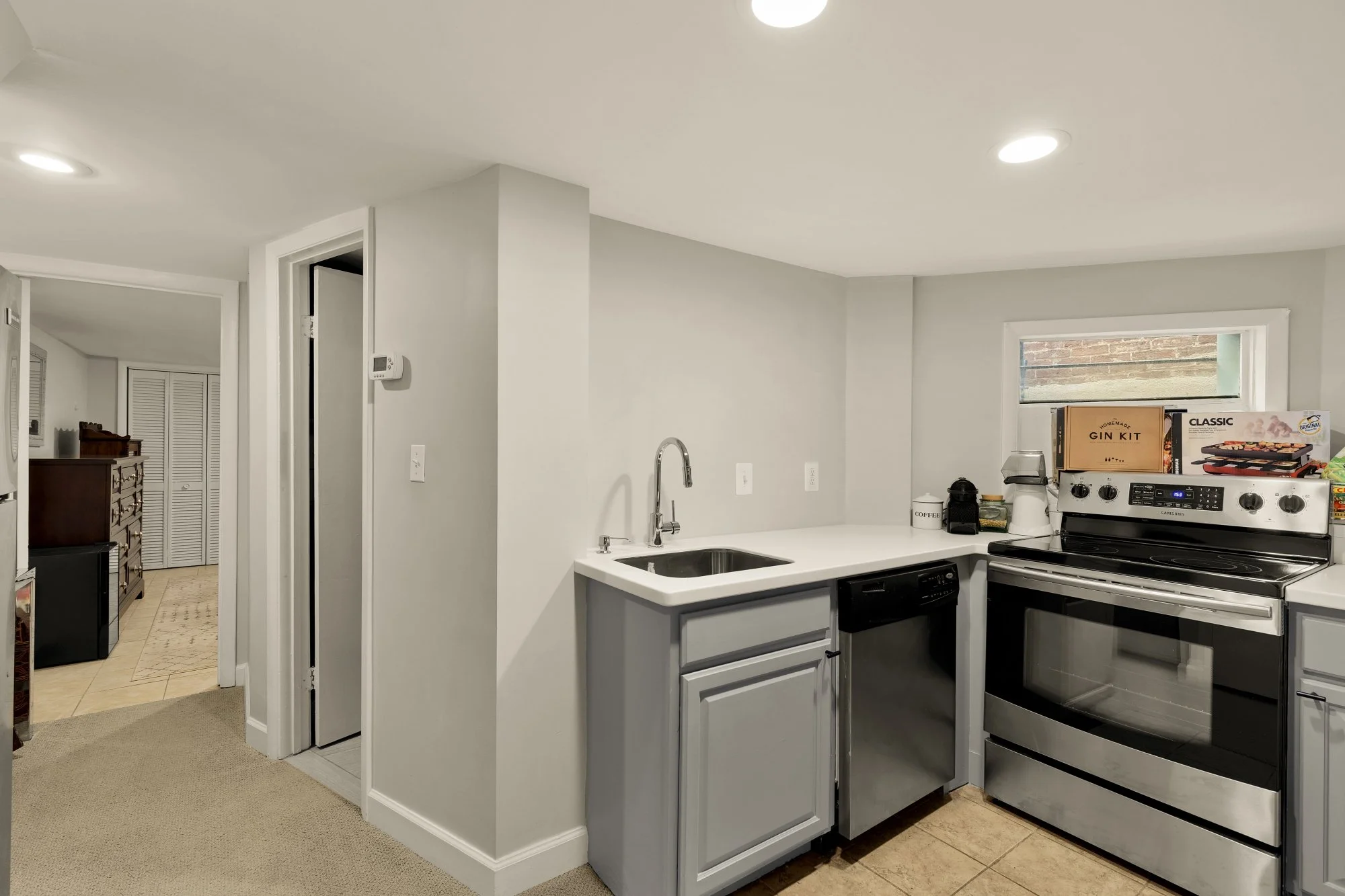 Modern kitchen with white cabinets, stainless steel stove, and a small window above the sink. Countertop has a "Homemade Gin Kit" box and a coffee maker. Adjacent room visible through doorway with a wooden dresser and beige carpet.