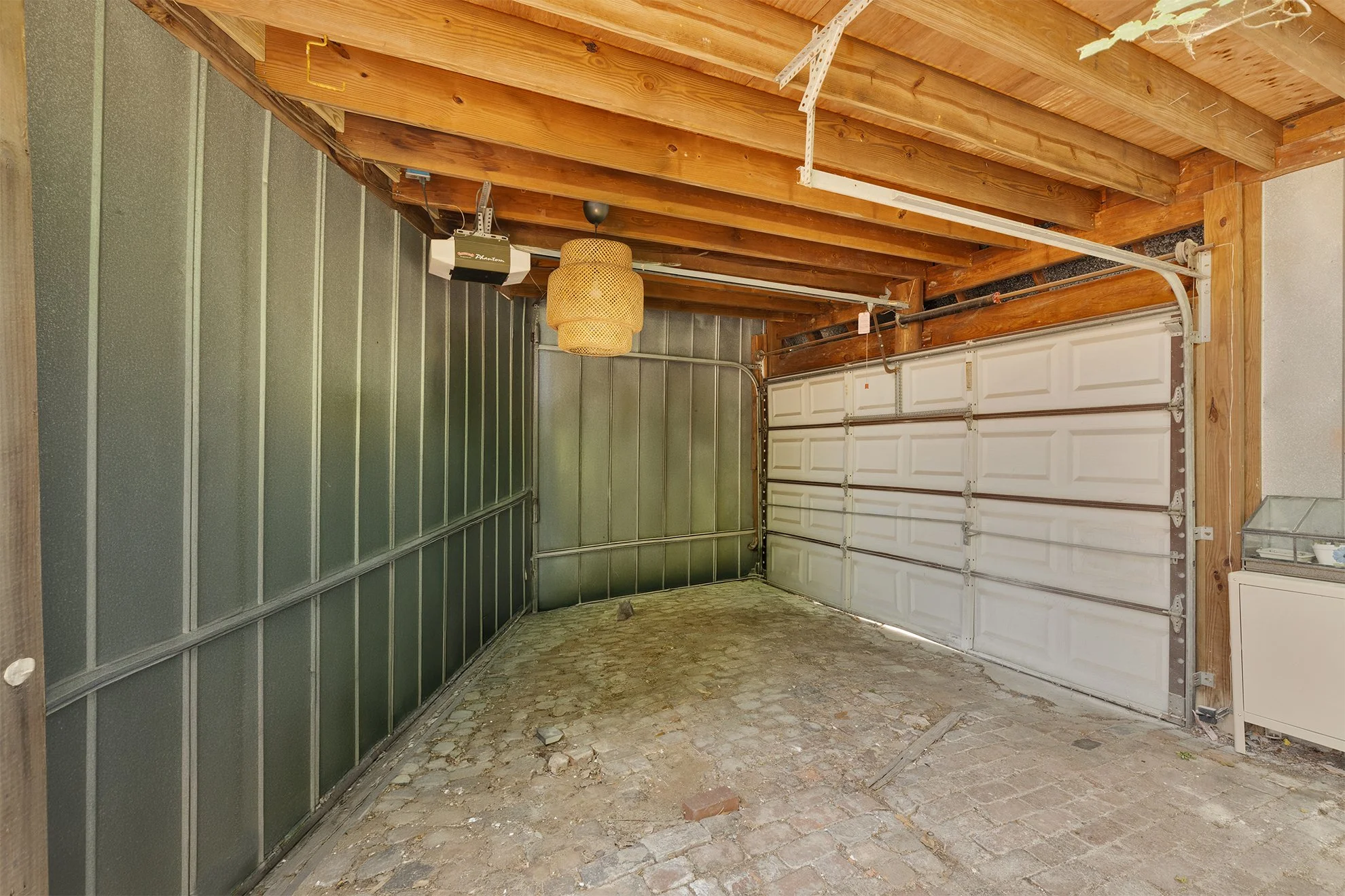 Empty garage with a closed white garage door, a cobblestone floor, and exposed wooden beams on the ceiling. A hanging light fixture and some electrical equipment are visible.