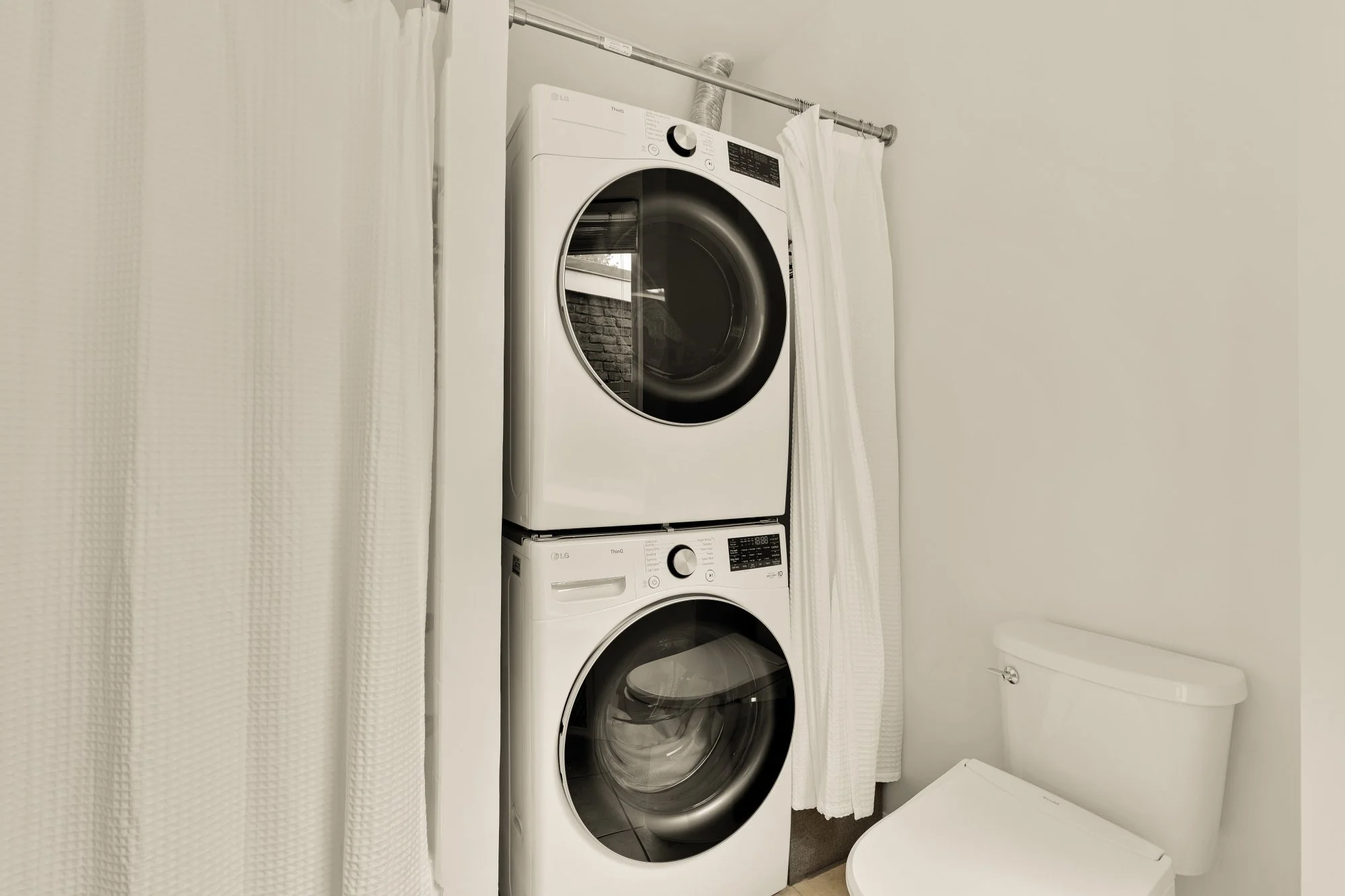A stacked washer and dryer in a laundry room, with white curtains on either side and a white toilet to the right.