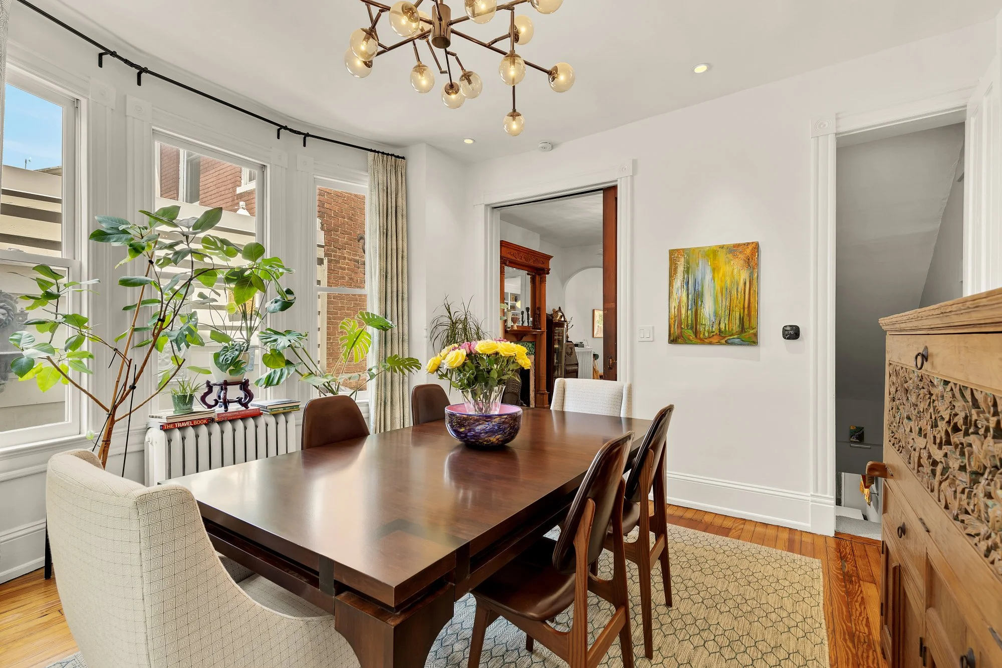 A dining room with a dark wood table and various chairs, a large potted plant near the window, a colorful flower arrangement, a painting of trees on the wall, a chandelier with multiple bulbs, and a view of the outside brick building through the wind
