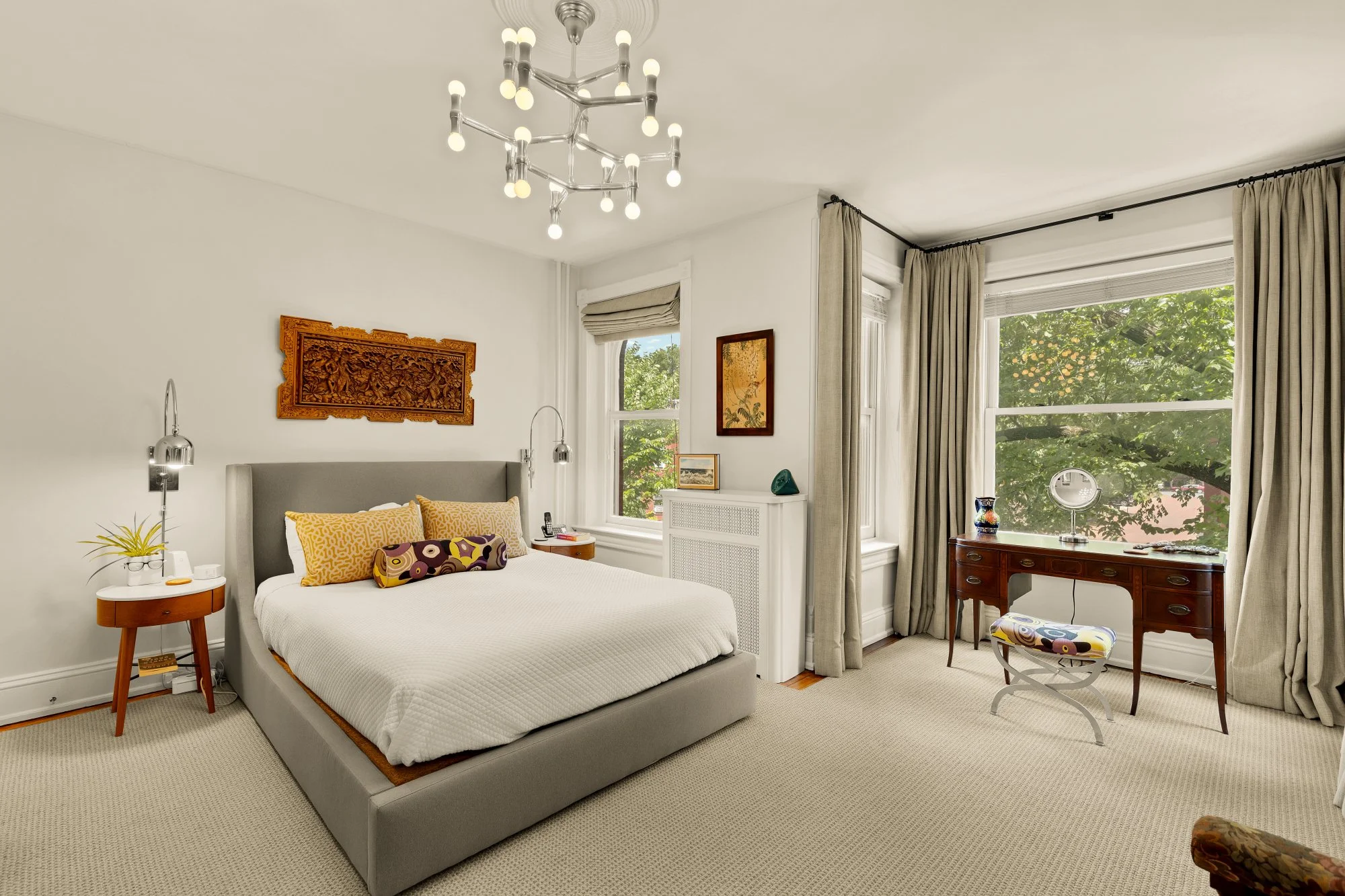 A bedroom with a gray bed and colorful pillows, wooden side table, modern chandelier, large windows with curtains, a decorative wall piece, and a vintage wooden desk with a chair, green vase, and a mirror.