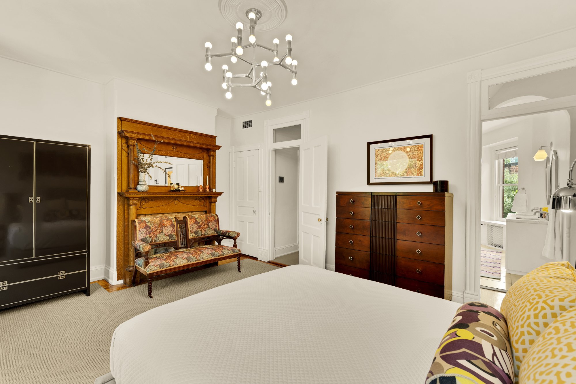A bedroom with a white bed, yellow and patterned pillows, a wooden dresser, a framed artwork, a vintage chair with floral upholstery, a black wardrobe, and a modern chandelier with LED bulbs on the ceiling.