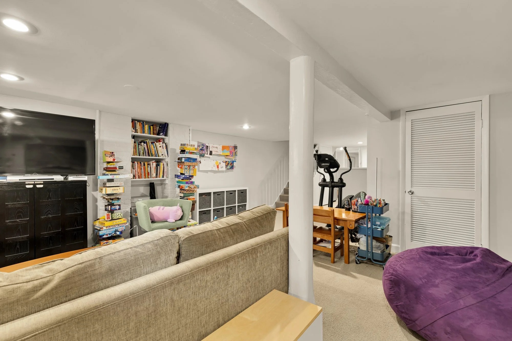 A cozy living space featuring a beige sofa, purple bean bag, bookshelf with books, a TV, children’s artwork on the wall, a small desk with art supplies, workout equipment, and a white door with a ventilation louver.