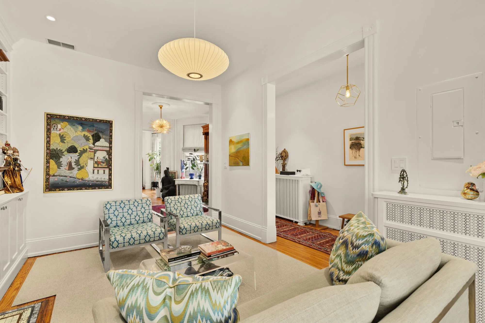Bright living room with white walls, wooden floors, patterned chairs, artwork, and various decorative items.