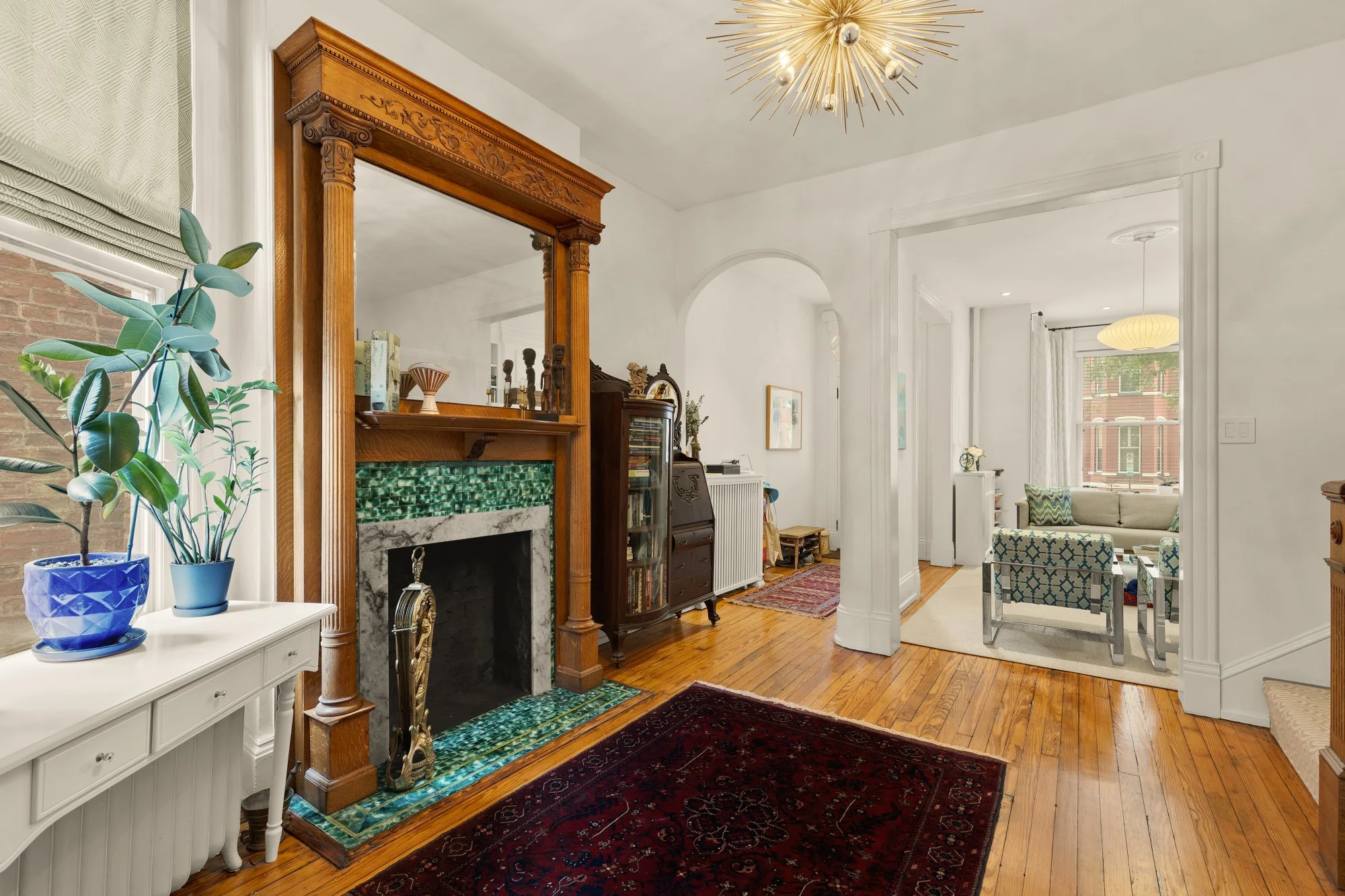 Interior view of a living room with a fireplace, decorated with vintage furniture, potted plants, and a colorful area rug, with a doorway leading to another sitting area