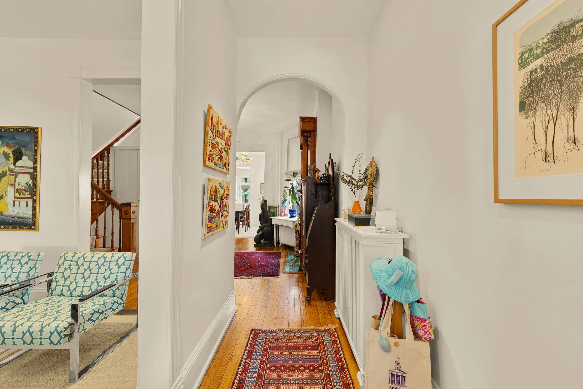 A hallway in a house with white walls, wooden flooring, and various artworks and decorations. There are chairs with patterned upholstery and a rug on the floor. The hallway leads to a dining area with a table and chairs visible in the background. Ite