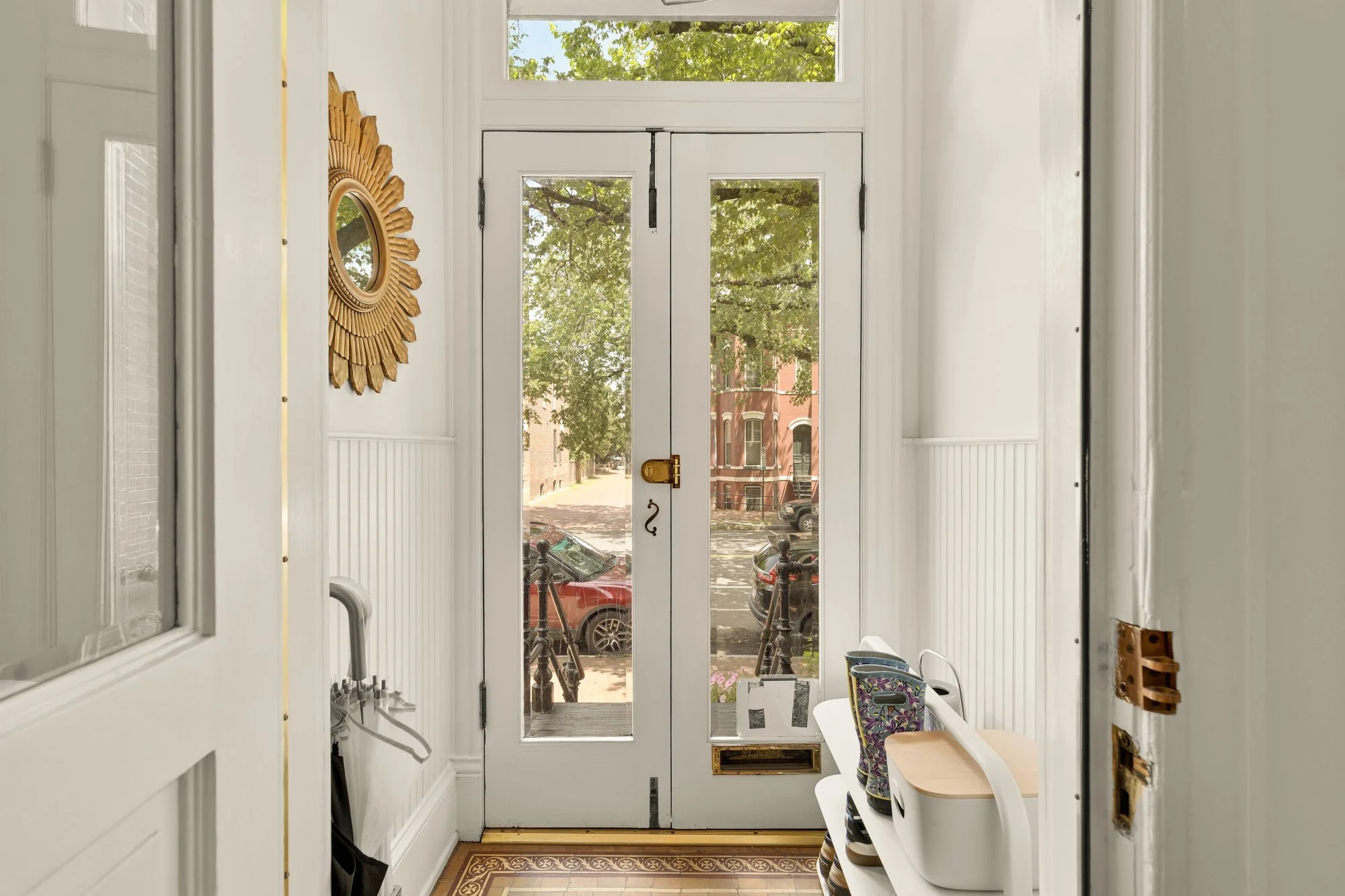Interior view of a white entryway with a glass-paneled door leading outside, visible cars parked on the street, a mirror with a sunburst frame on the wall, and a chair with a floral-patterned bag on it.