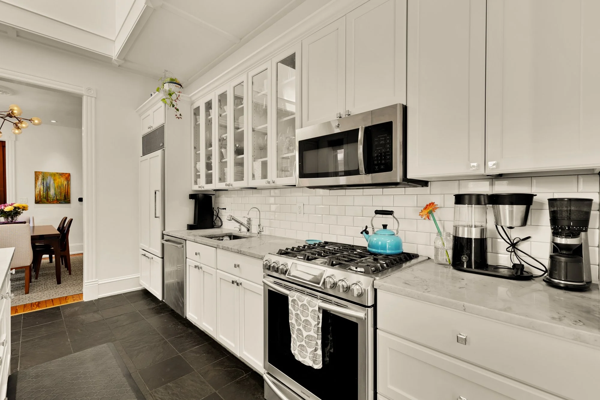Kitchen with white cabinets, stainless steel microwave, stove, and sink. Black tile floor with a gray mat, and various small appliances on the counter, including a blue kettle and coffee machines.