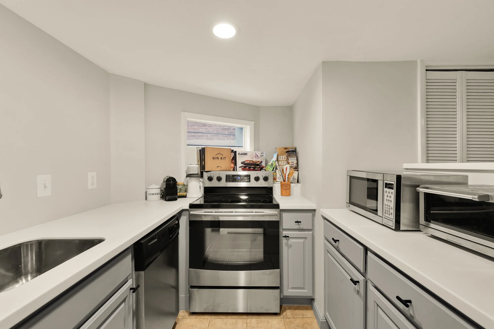 Small kitchen with white counters and cabinets, stainless steel appliances including a stove, microwave, and dishwasher, black kitchen gadgets, and a window with items on the sill.