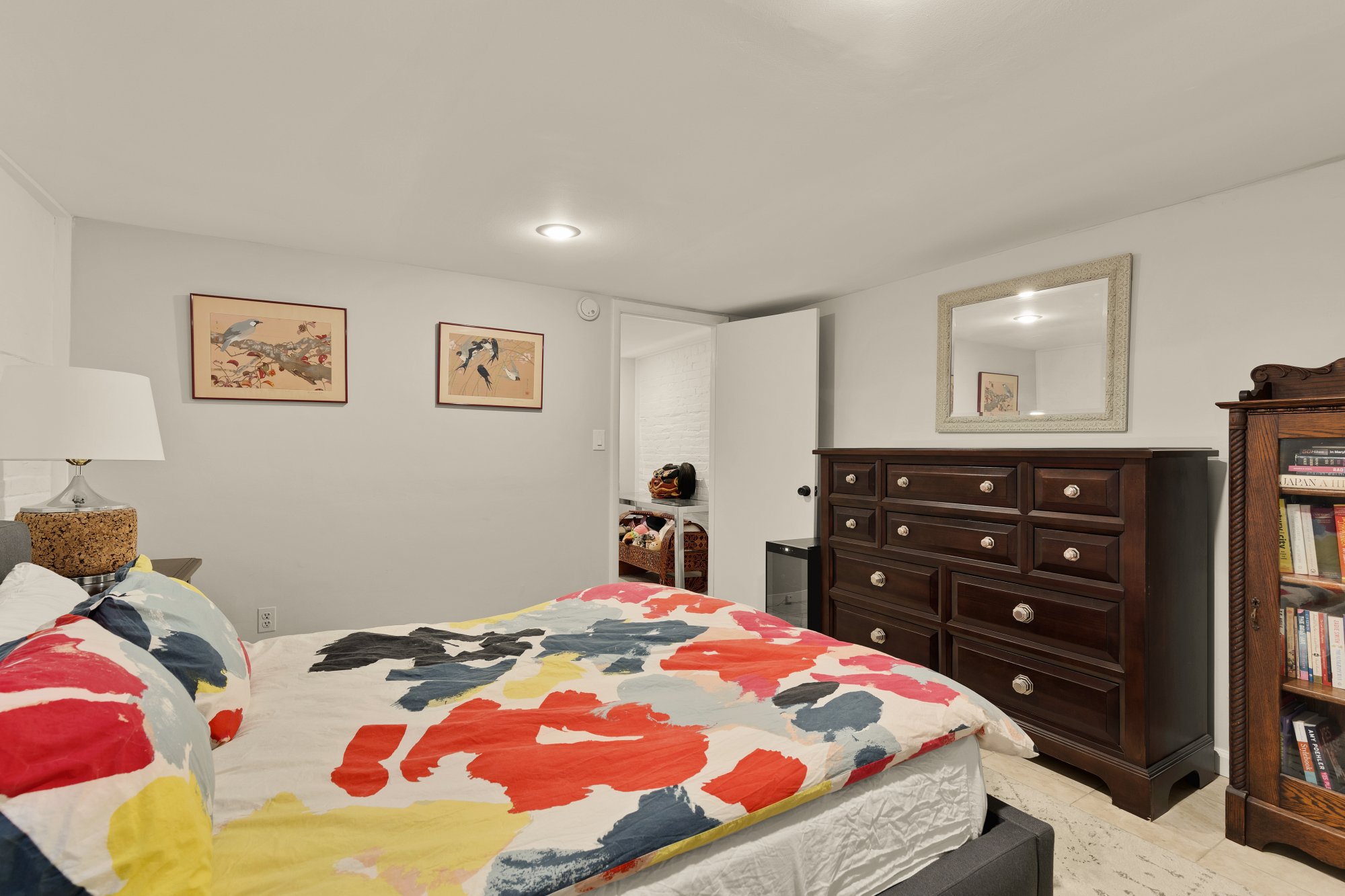 A bedroom with a bed that has a colorful abstract pattern comforter and pillows. There are two framed bird-themed art pieces on the wall, a white lamp on a nightstand, a dark wooden dresser with a mirror above, and a piece of furniture with books on 