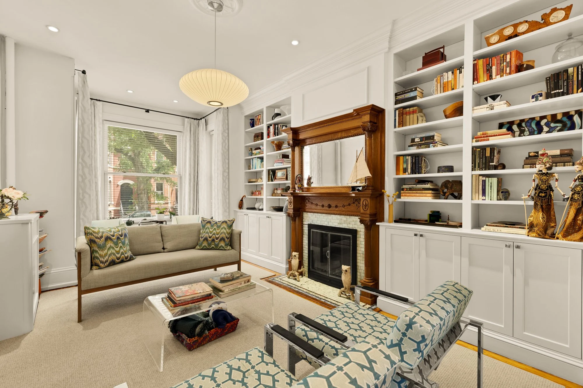 Living room with white walls, built-in shelves, a fireplace with a wooden mantle, beige sofa with colorful patterned pillows, a glass-top coffee table with magazines, and windows with white curtains.