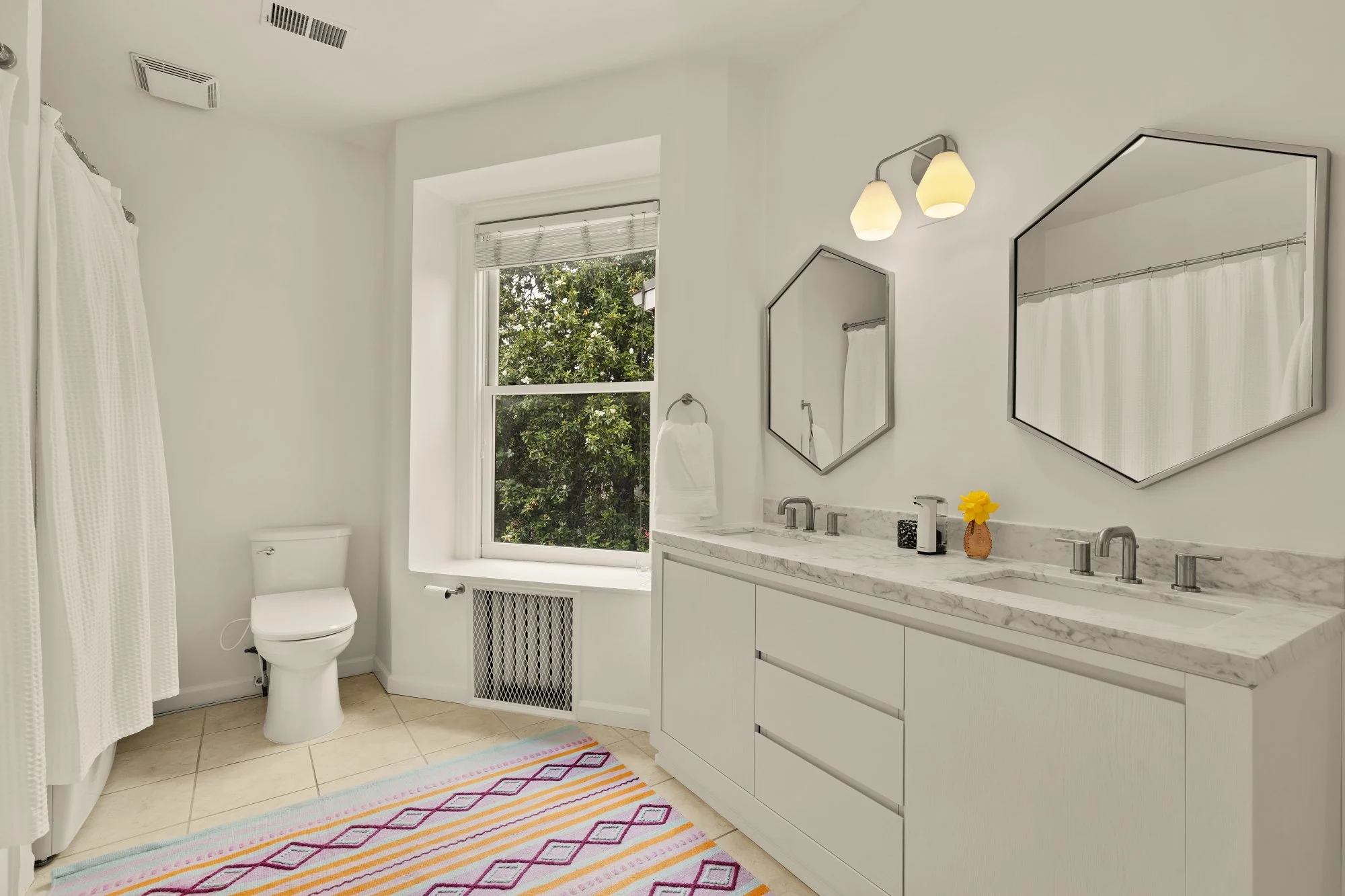 Bright bathroom with a window, white vanity with marble countertop, two mirrors, light fixture, and decorative items including a yellow flower, with a colorful rug on beige tile floor.