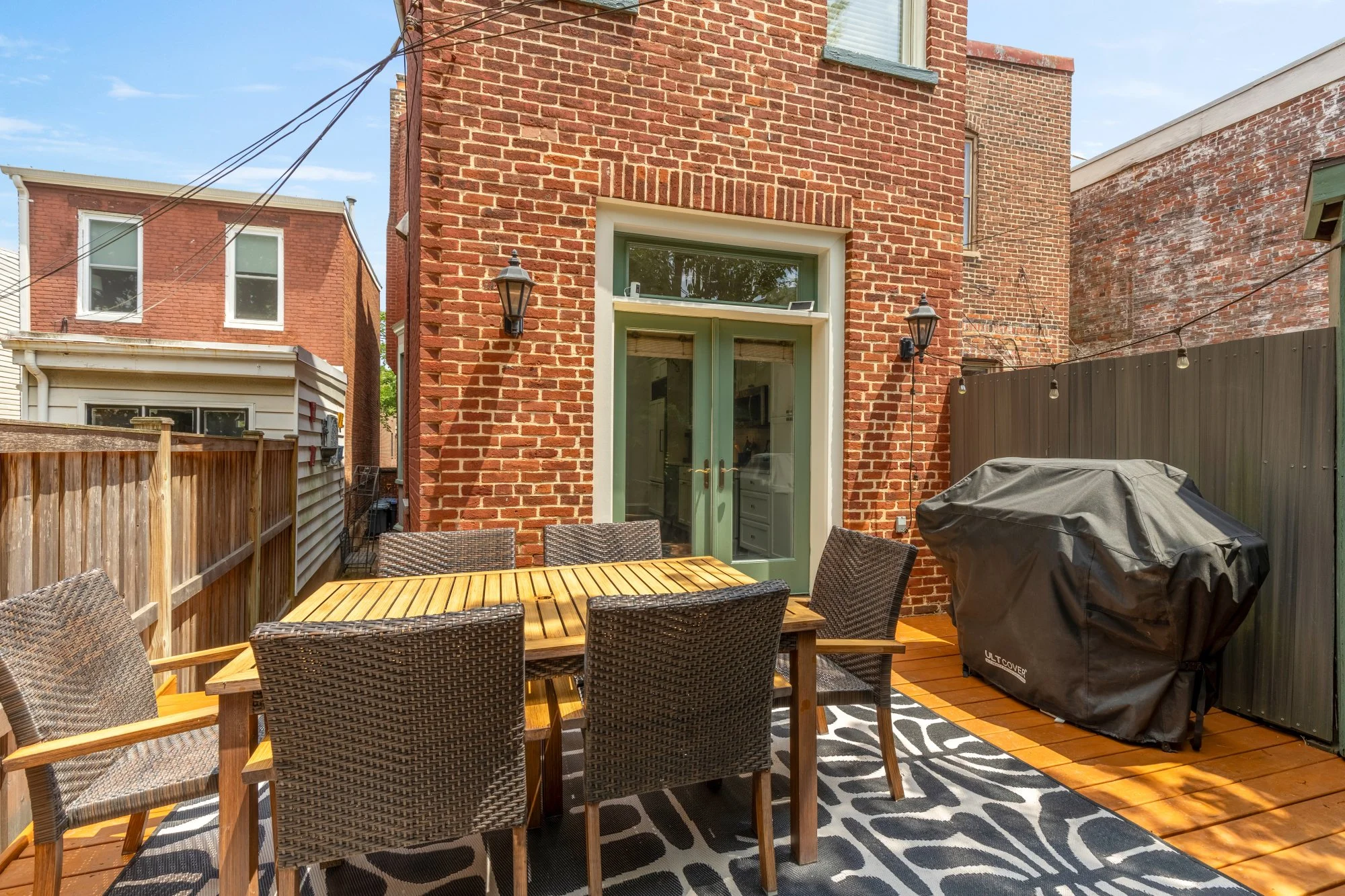 A backyard patio with a rectangular wooden dining table and six wicker chairs. A black barbecue grill covers part of the deck. The back door is green framed with white trim. The porch is enclosed with wood fences and string lights hang across the yar