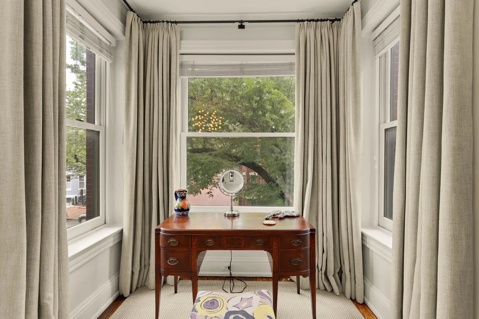 A sunroom with large windows showing trees and a brick building outside. Inside, there is a wooden desk with a decorative vase, a mirror, and some small objects. Cream-colored curtains frame the windows, and a cushioned stool with a colorful pattern 
