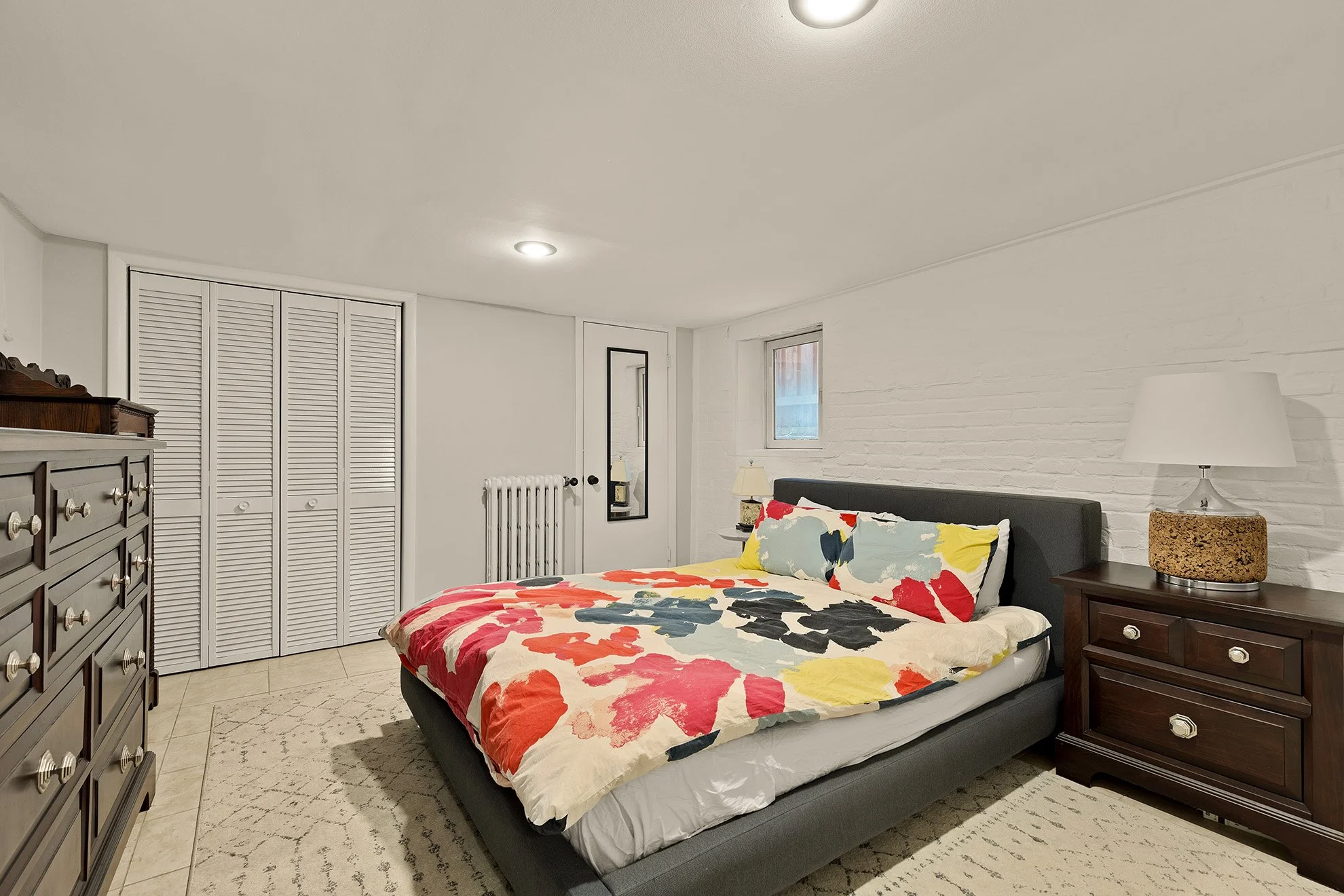 A bedroom with a large bed covered in a colorful floral comforter, dark wood nightstands, white walls with a brick texture, a window, closet with louvered doors, and a dresser, with a patterned rug on light-colored tile floor.