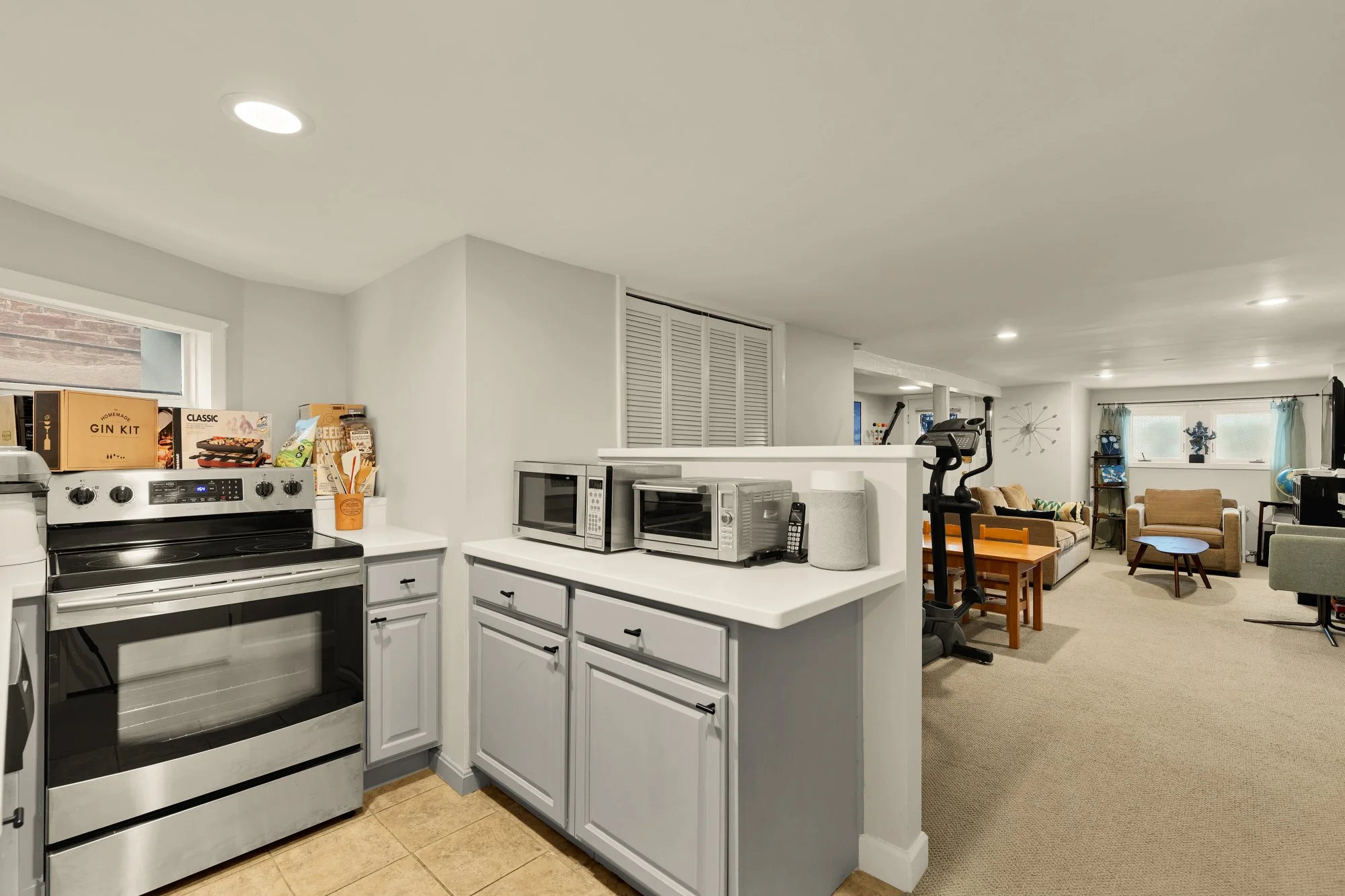 View of a kitchen and living room area with white cabinets, a stove, microwaves, and appliances in the kitchen; and couches, a bookshelf, and a window in the living room, with neutral-colored walls and carpeted flooring.