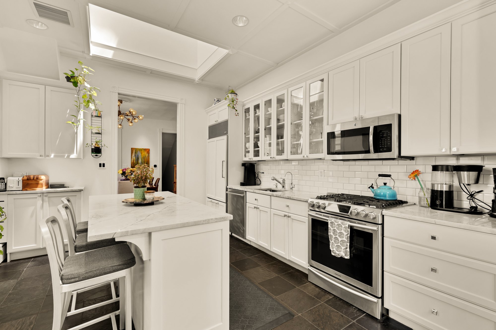A bright, modern kitchen with white cabinets, marble countertops, and a stainless steel gas stove. There is a kitchen island with seating, potted plants, and various small appliances including a coffee maker and toaster.