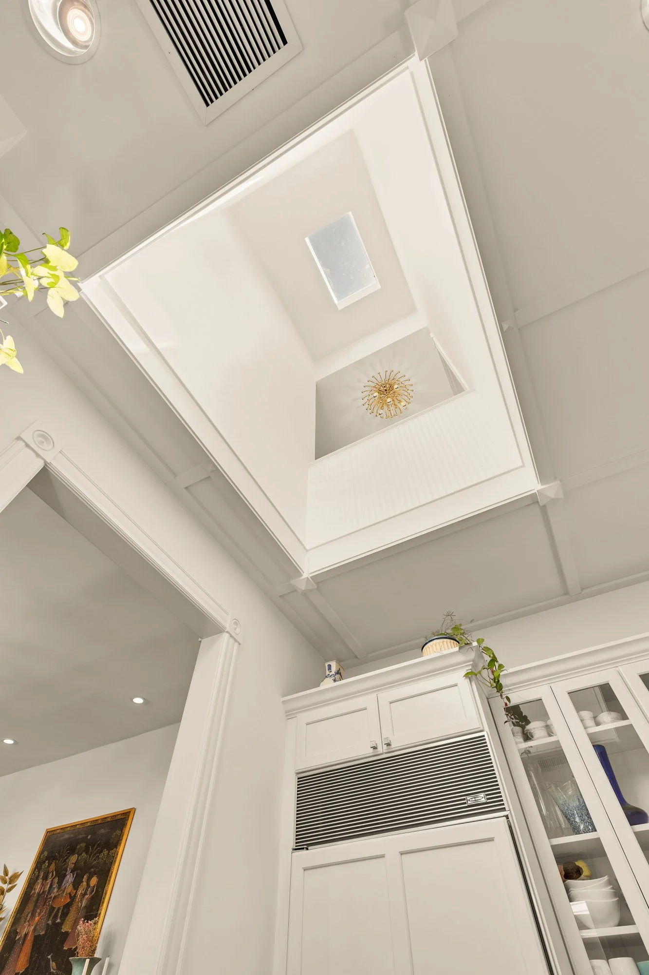 Ceiling view of a white kitchen with skylights, a gold chandelier, and cabinets with a built-in microwave, decorated with plants and artwork.