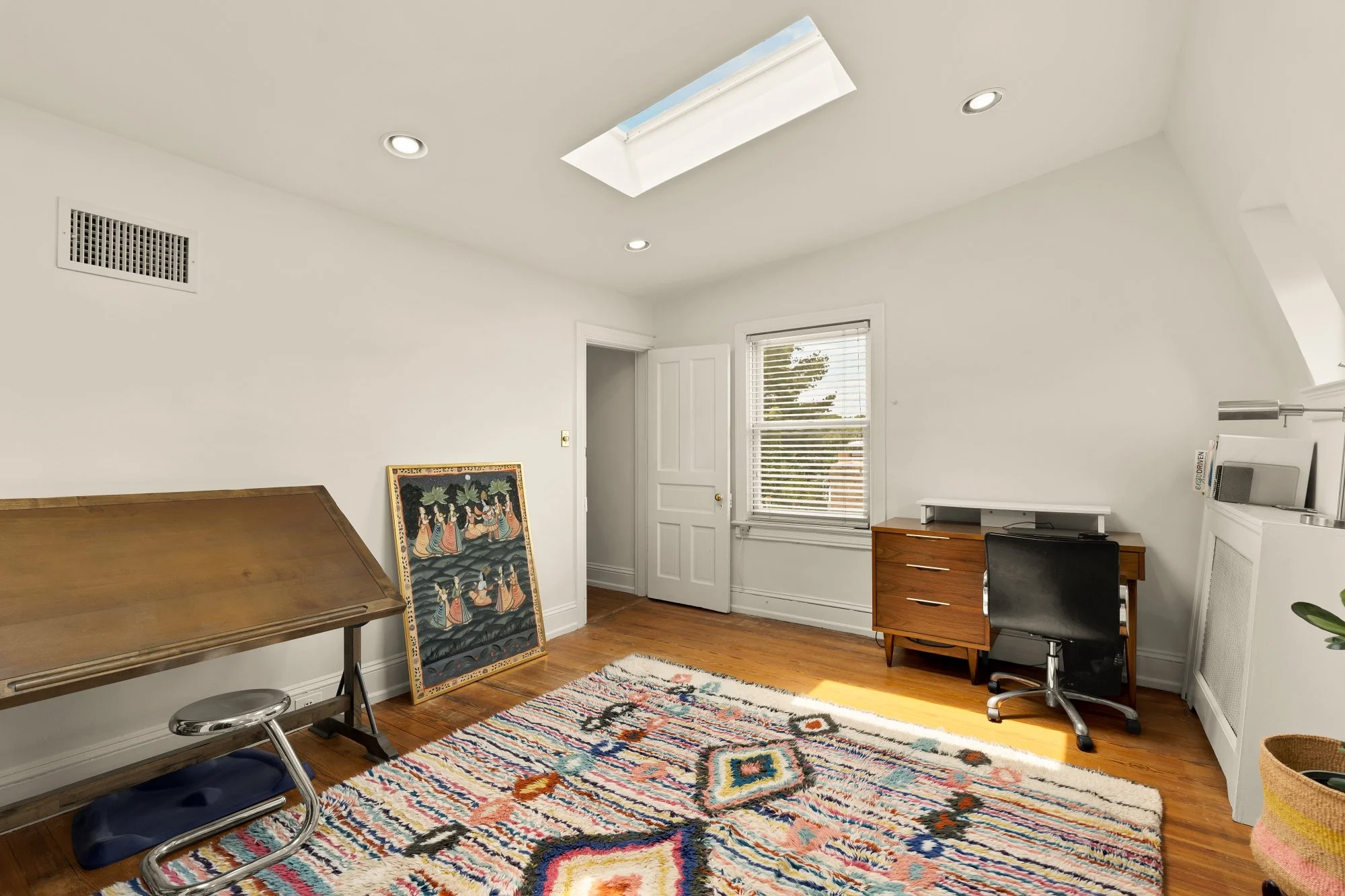 A room with a colorful patterned rug on hardwood floor, white walls, a wooden desk with a chair, large window with blinds, a door, framed artwork leaning against the wall, and a white cabinet with a desktop computer on top.