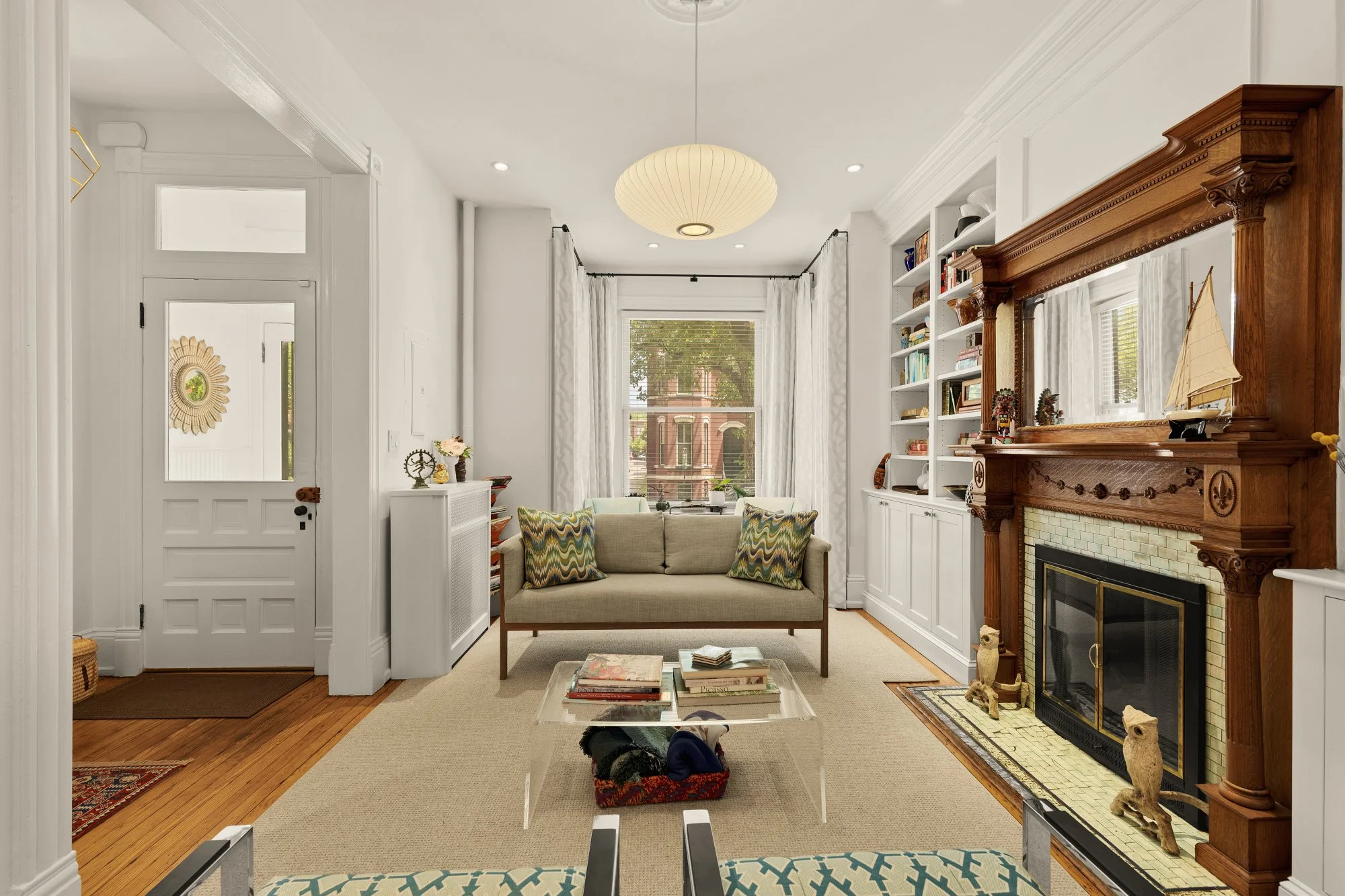 Living room with a beige sofa, armchairs, built-in bookshelves, a fireplace with decorative mantel, large window with white curtains, and a ceiling light fixture.