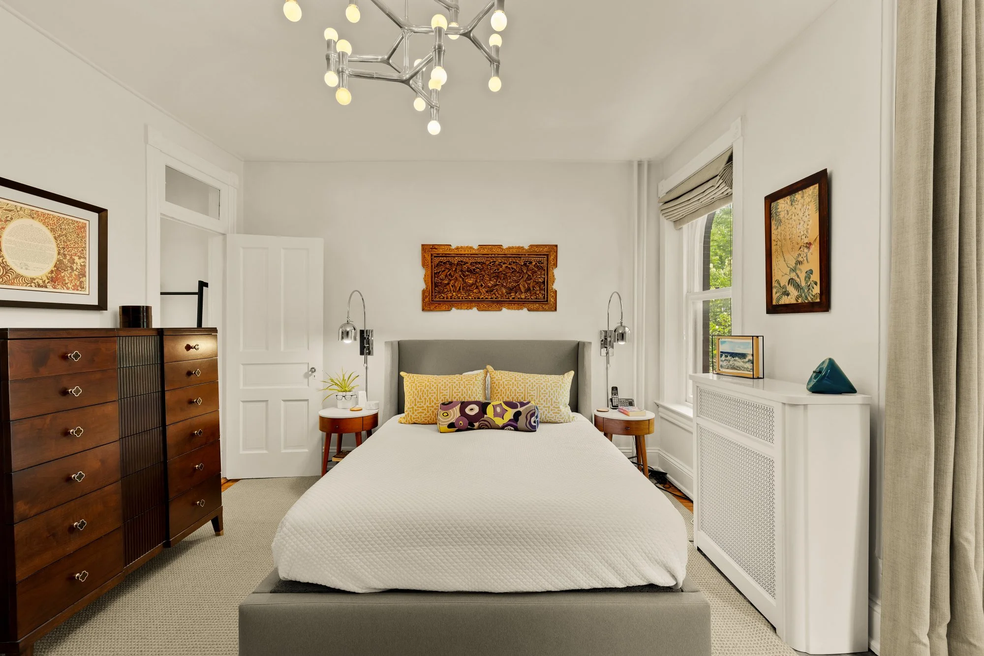 A bedroom with a bed in the center, a wooden dresser to the left, and a white radiator cover to the right. The bed has yellow and purple patterned pillows, and there are two small side tables with lamps beside it. The room has white walls, a decorati