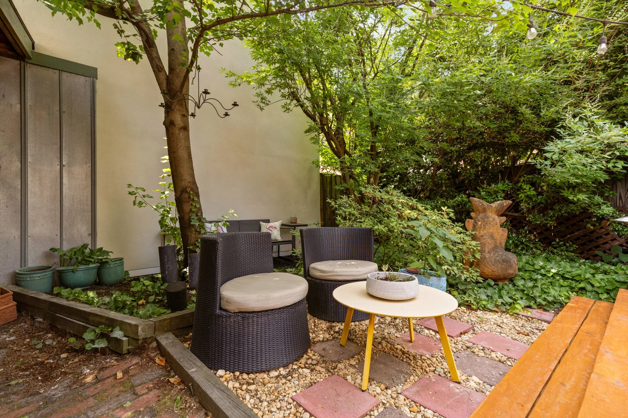 A cozy backyard patio with black wicker chairs with white cushions and a yellow table, surrounded by lots of green foliage, a tree, and hanging string lights.