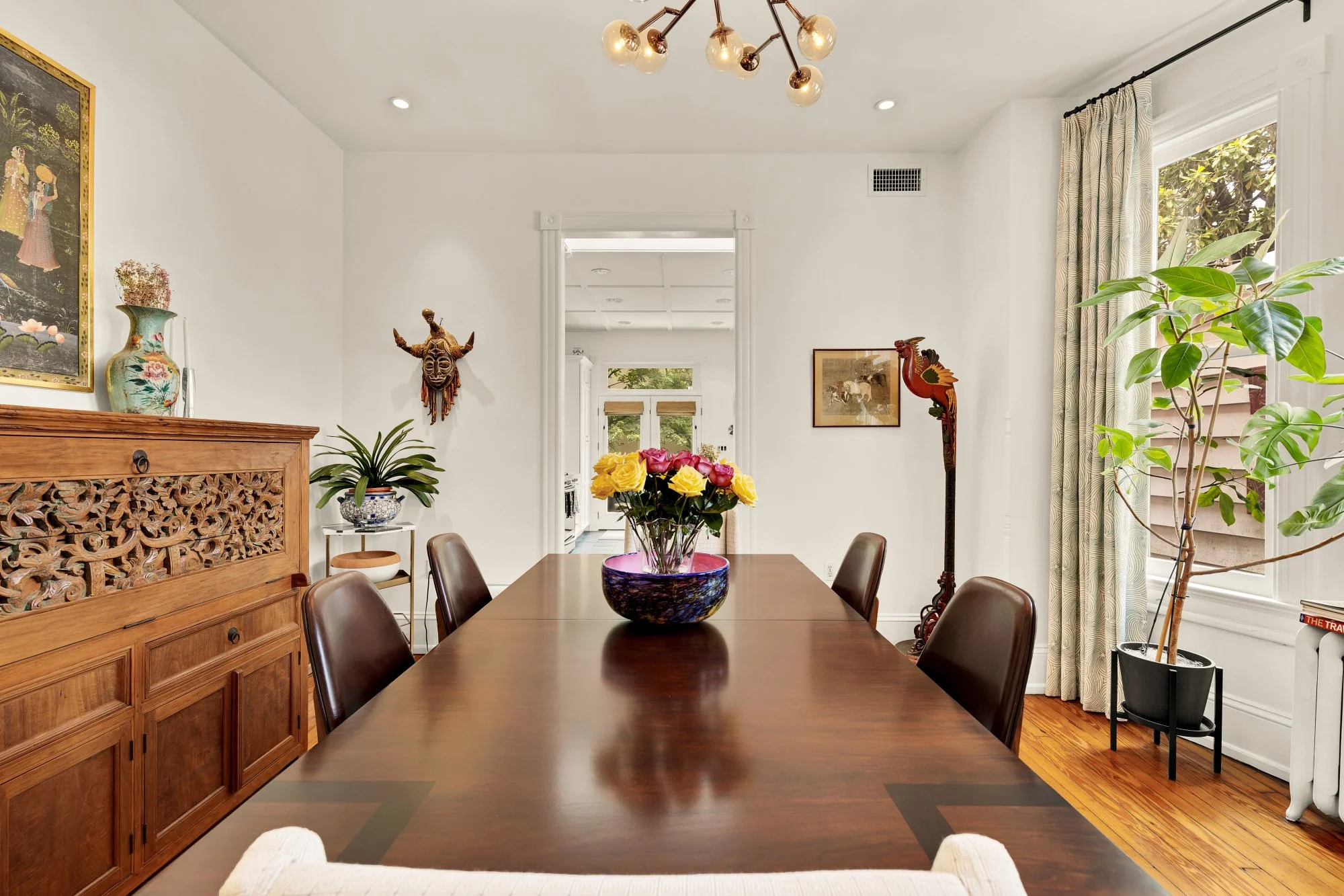 A dining room with a wooden table and chairs, a large flower arrangement in a blue bowl, a decorative wooden sideboard, framed artwork, a colorful tribal mask, and various plants near windows with patterned curtains.