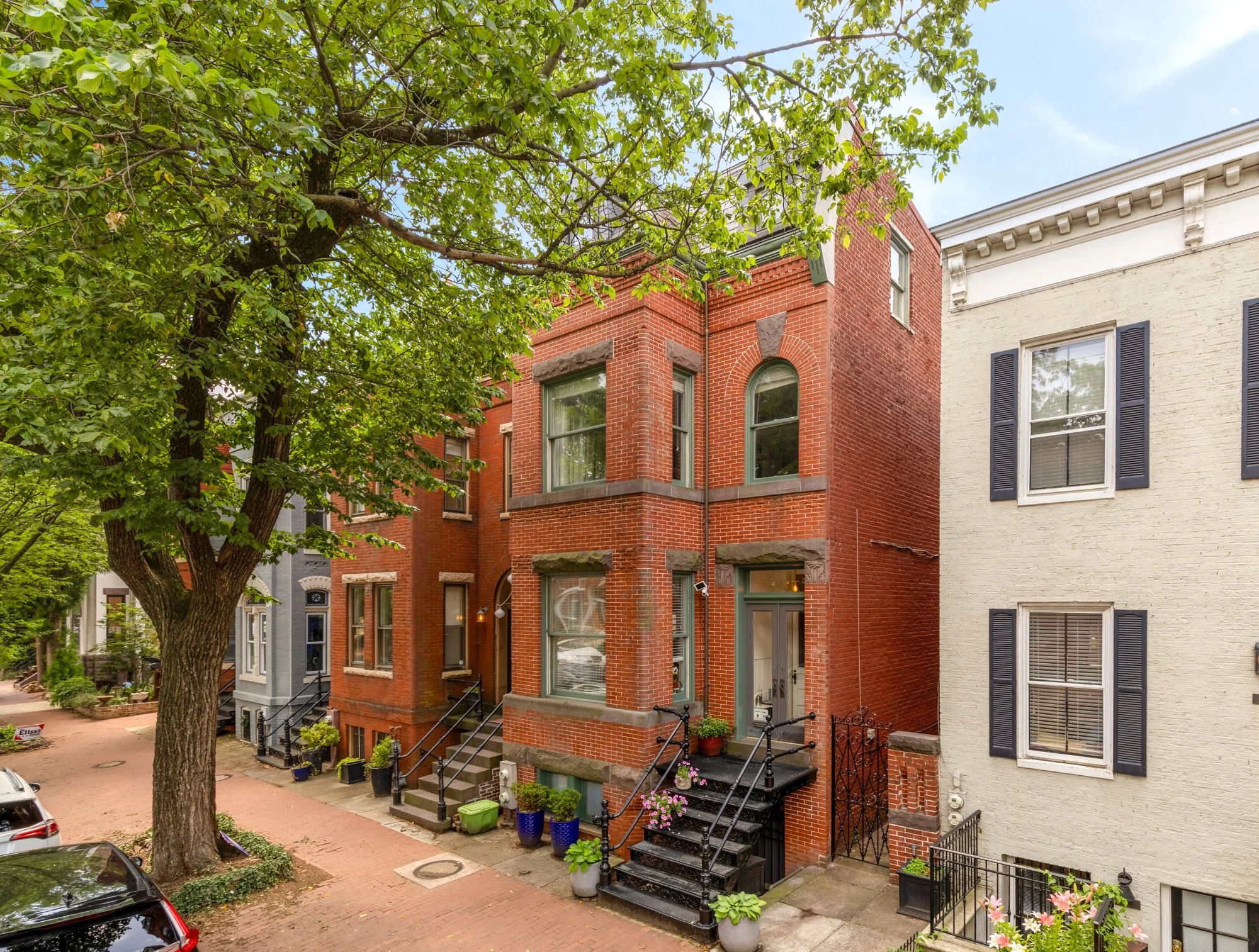 Row of historic brick townhouses along a brick-paved sidewalk with potted plants, bicycles, and large leafy trees.