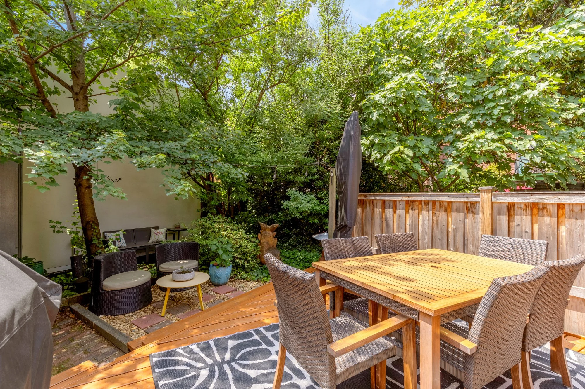 A backyard patio with a wooden dining table surrounded by wicker chairs, a small seating area with cushions, potted plants, a large tree with green leaves, a wooden fence, and a black patio umbrella.