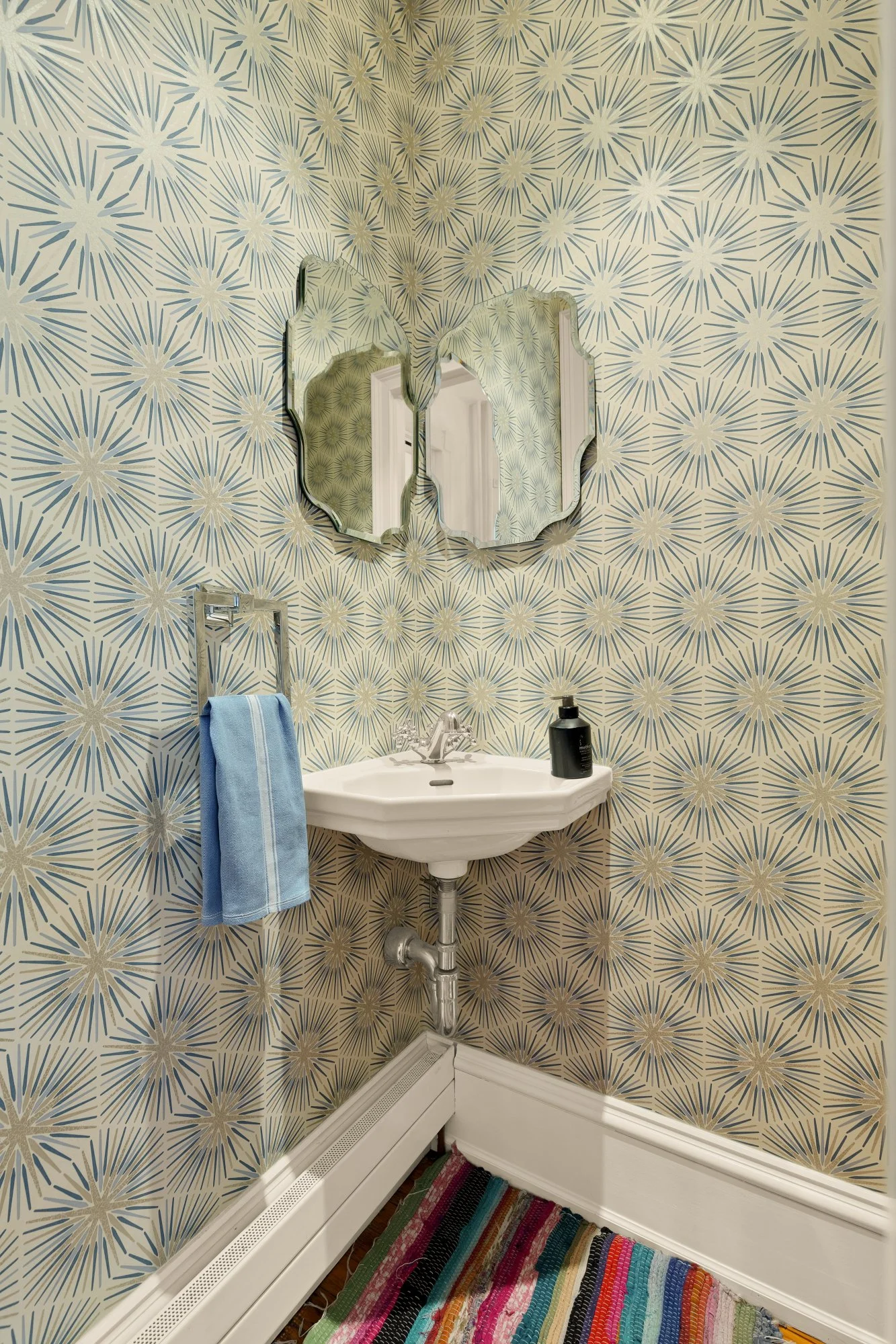 Small bathroom corner with a white sink, a black soap dispenser, a towel rack with a blue towel, a colorful striped rug, patterned wallpaper with starburst design, and a decorative wall mirror.