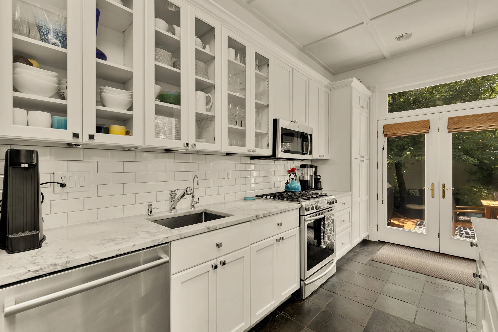 Kitchen with white cabinets, marble countertops, white subway tile backsplash, black stove, microwave, and various kitchen items on open shelves. Doors lead outside to a patio area with trees.