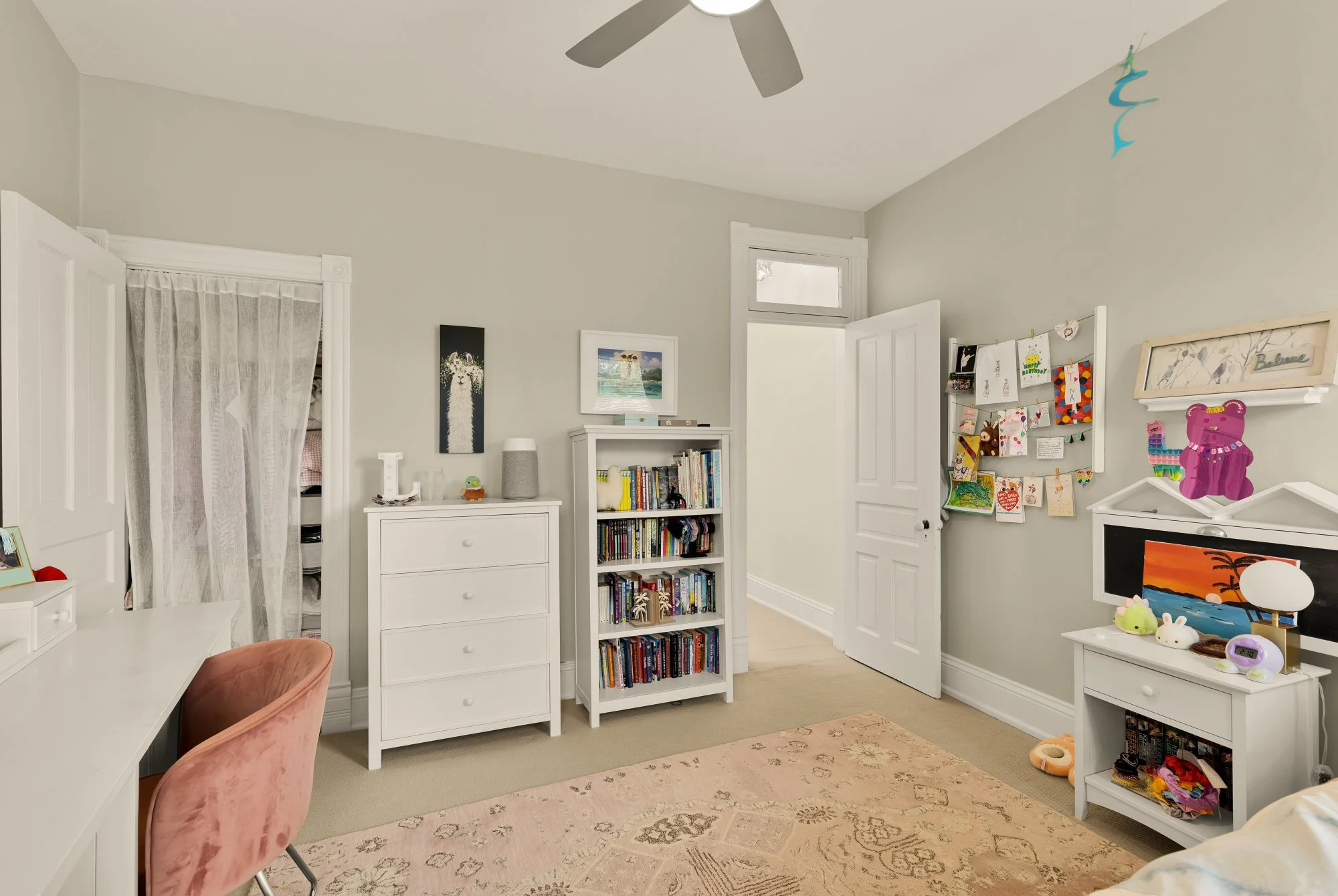 A cozy children's bedroom with white walls and a ceiling fan. Furniture includes white dressers, a bookshelf filled with books, a changing table, and a desk with a pink chair. Decorations include framed pictures, artwork, and crafts hanging on a bull