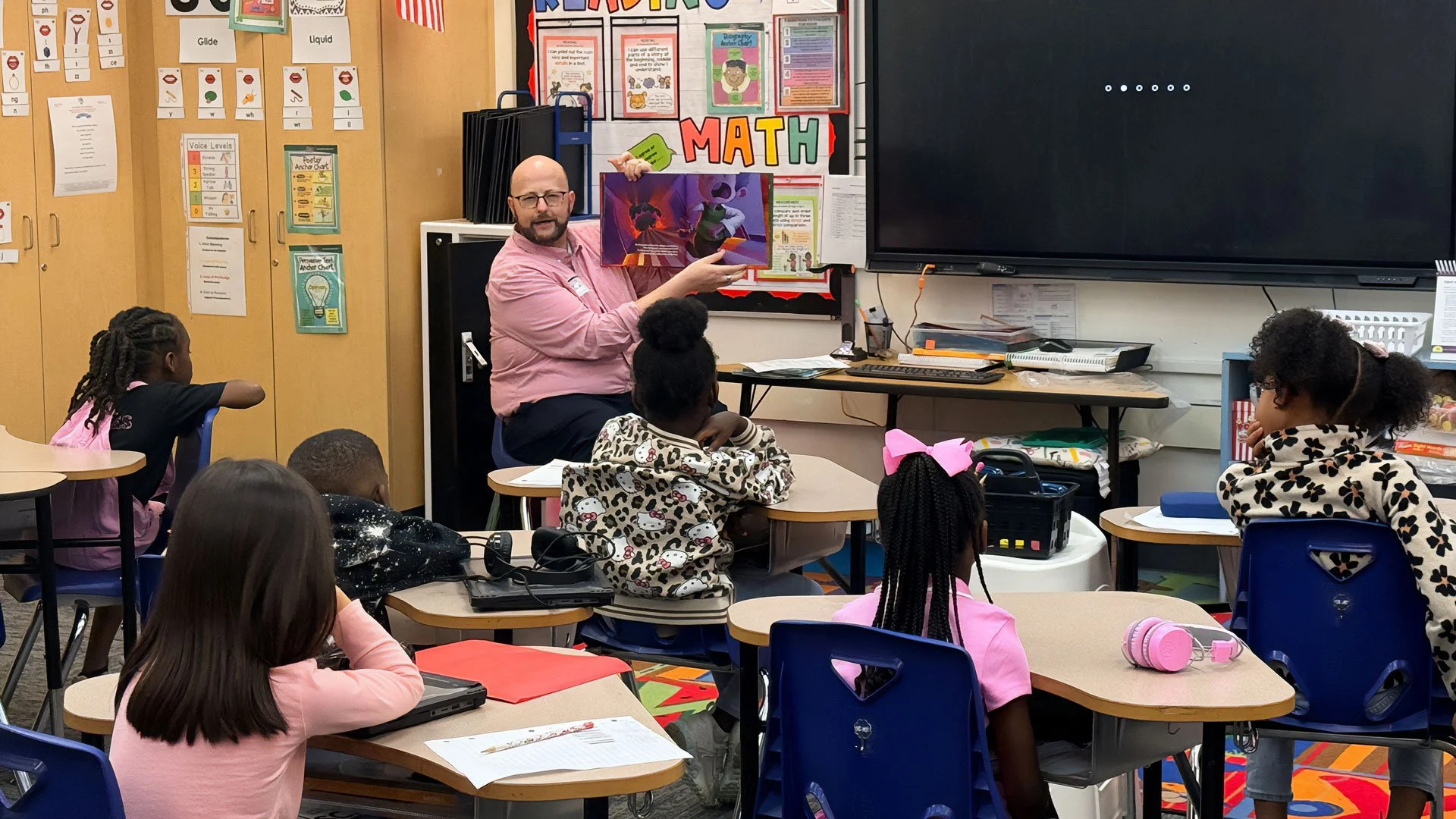 A teacher reads a picture book to students in a classroom, with children sitting at desks listening attentively. The classroom has colorful decorations, a blackboard, and educational posters on the walls.