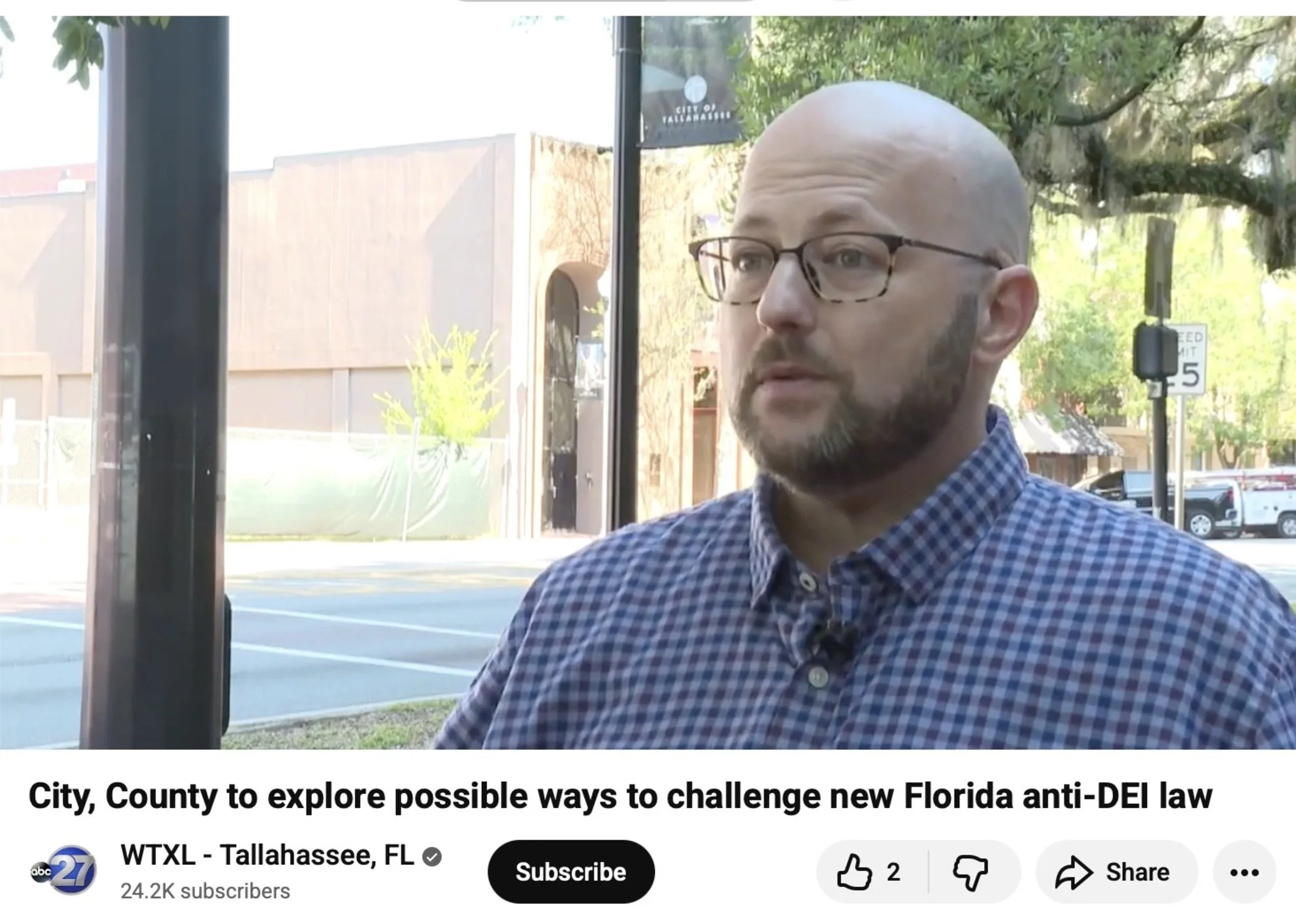 A man with glasses and a beard wearing a blue checkered shirt is sitting inside a bus or train near a large window. Outside, there are trees, a building, parked cars, and a street view in Tallahassee, Florida.