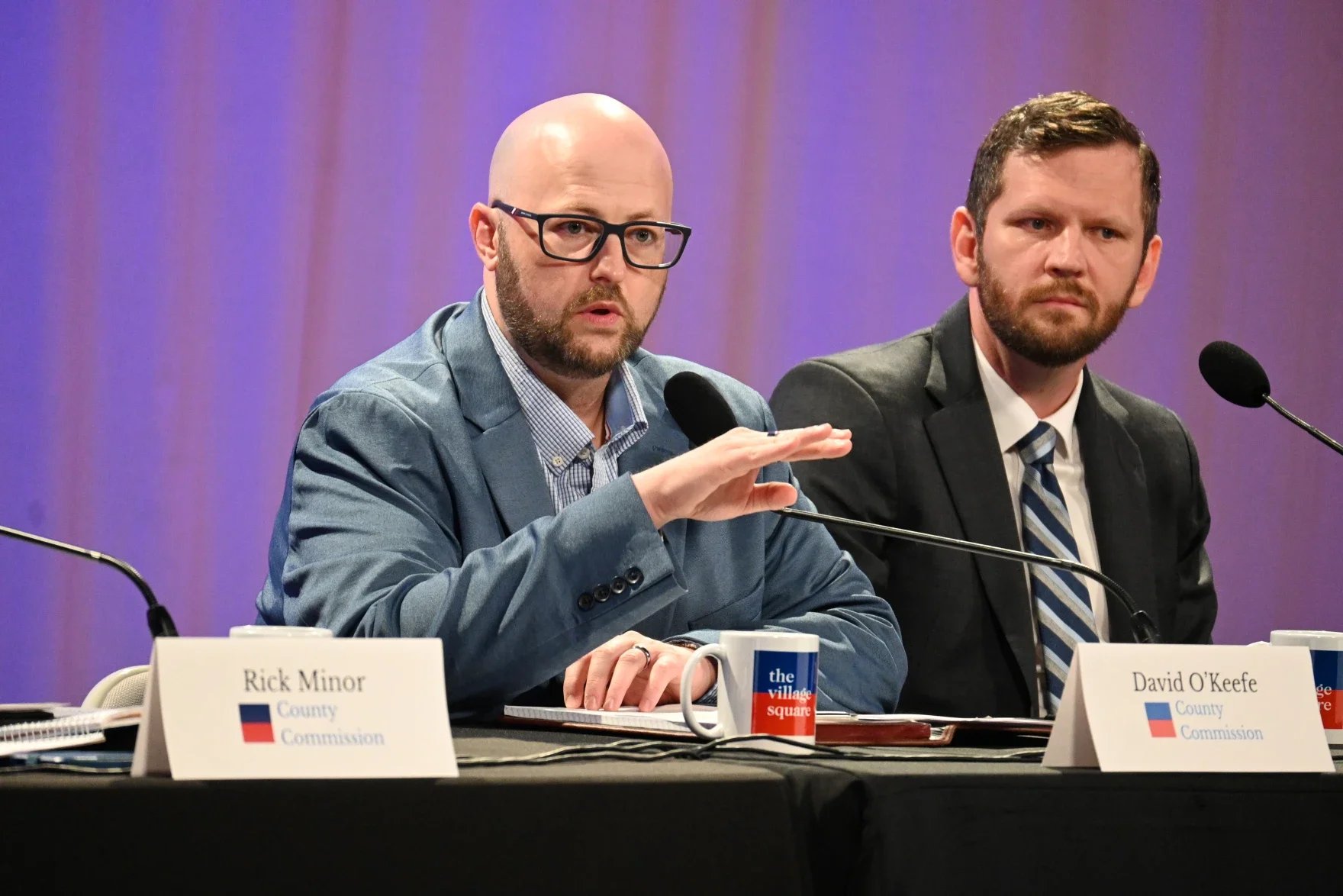 Two men sitting at a table speaking into microphones during a formal meeting, with name tags in front of them. The man on the left is bald with glasses, wearing a blue jacket, gesturing with his hand. The man on the right has dark hair and a beard, wearing a suit and tie. The table has a cup with a logo and several papers.