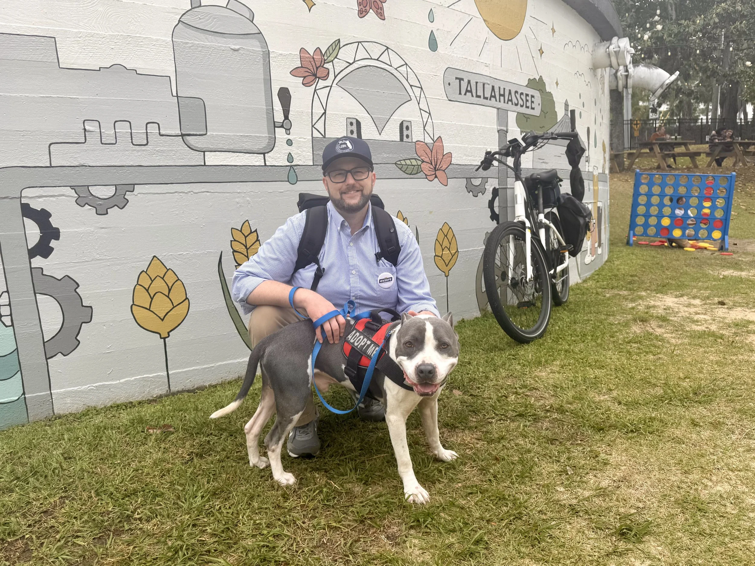 A man in glasses, a cap, and a button-up shirt kneels next to a happy black and white dog wearing a harness with the words 'Adopt Me'. They are outdoors with a painted wall behind them that features a mural including a water faucet, flowers, and a sign that reads 'TALLAHASSEE.' There is a bicycle and a large connect four game in the background, along with picnic tables and trees.