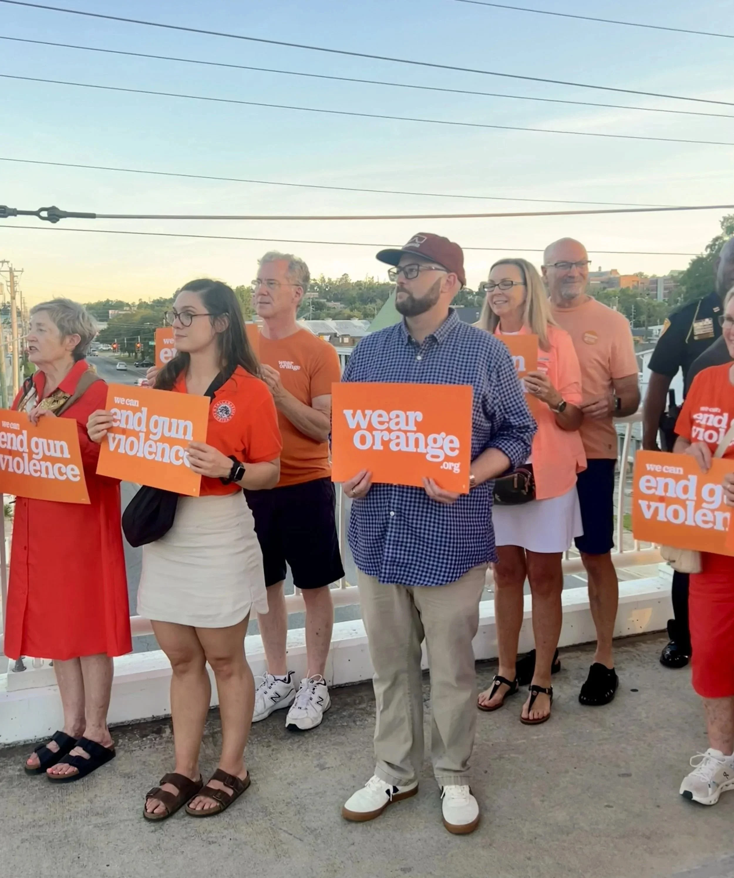 Group of diverse people standing outdoors holding orange signs that say "wear orange" and advocating to end gun violence, with a cityscape and power lines in the background during sunset.