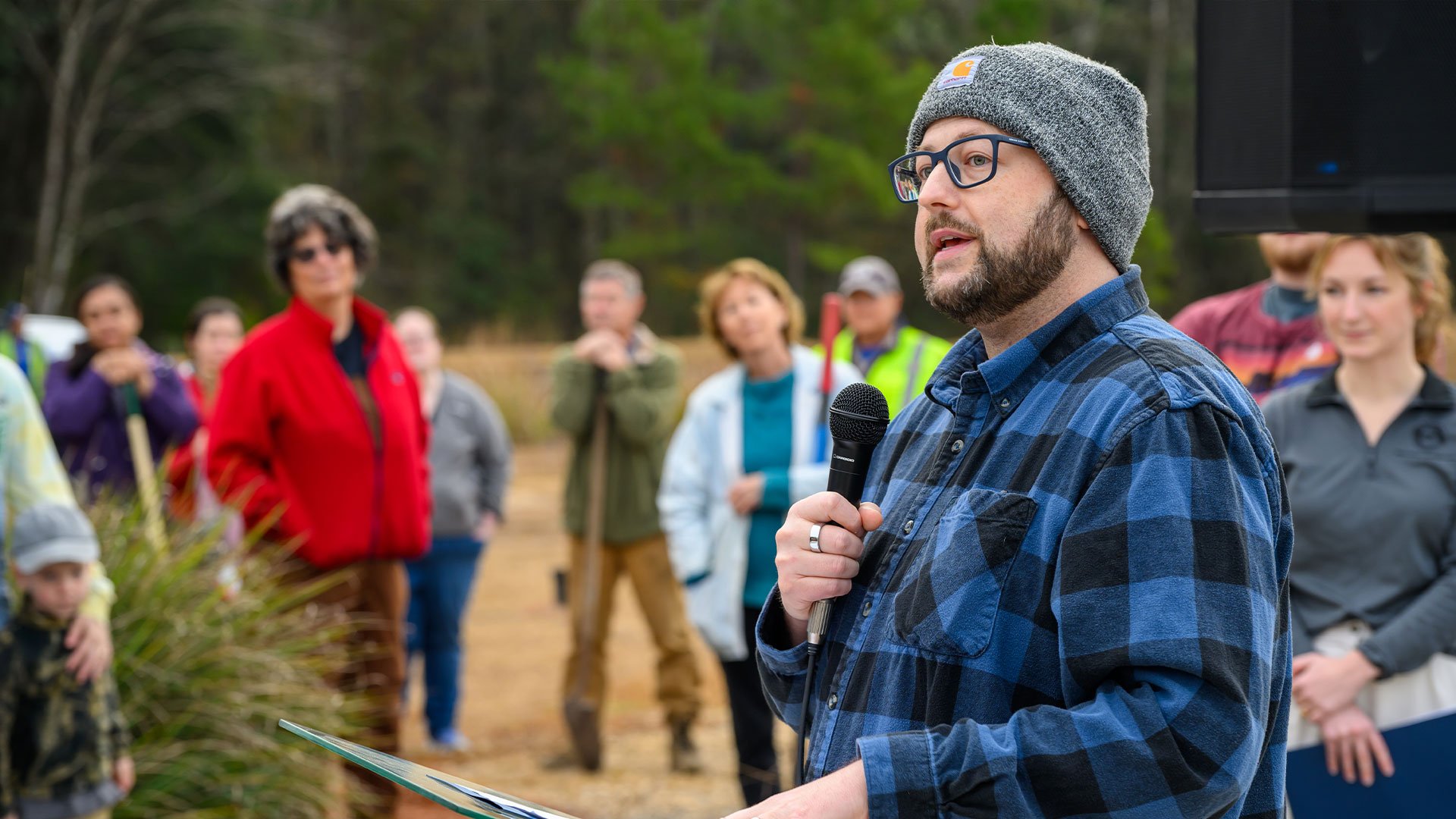 Man wearing glasses and a gray beanie speaks into a microphone at an outdoor event, with a group of people listening in a wooded area.