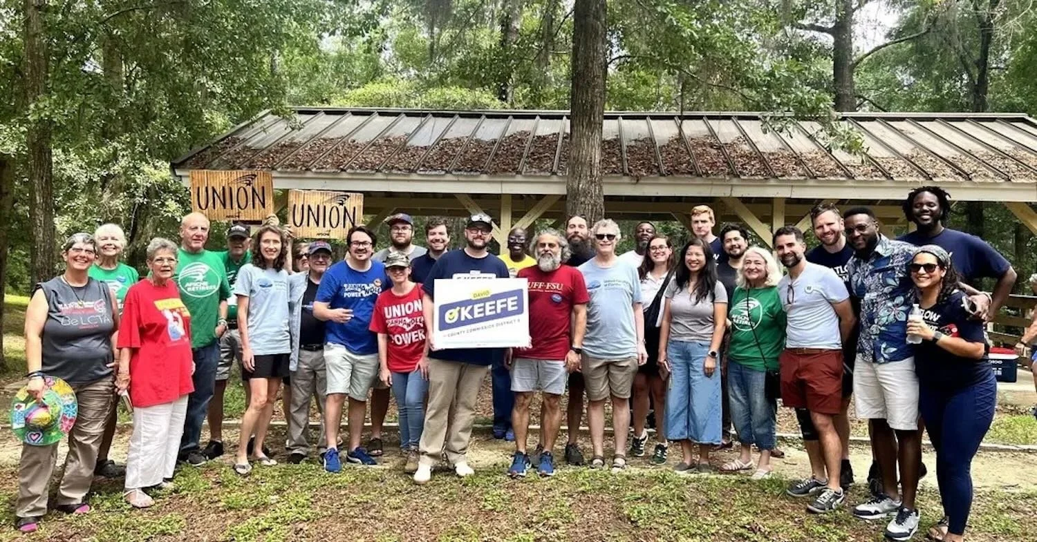 A group of people gathered outdoors for a photo, standing in front of a wooden structure with a metal roof in a wooded area. Some are holding signs that say 'Union.' The group appears to be diverse in age and ethnicity, and they are smiling at the camera.