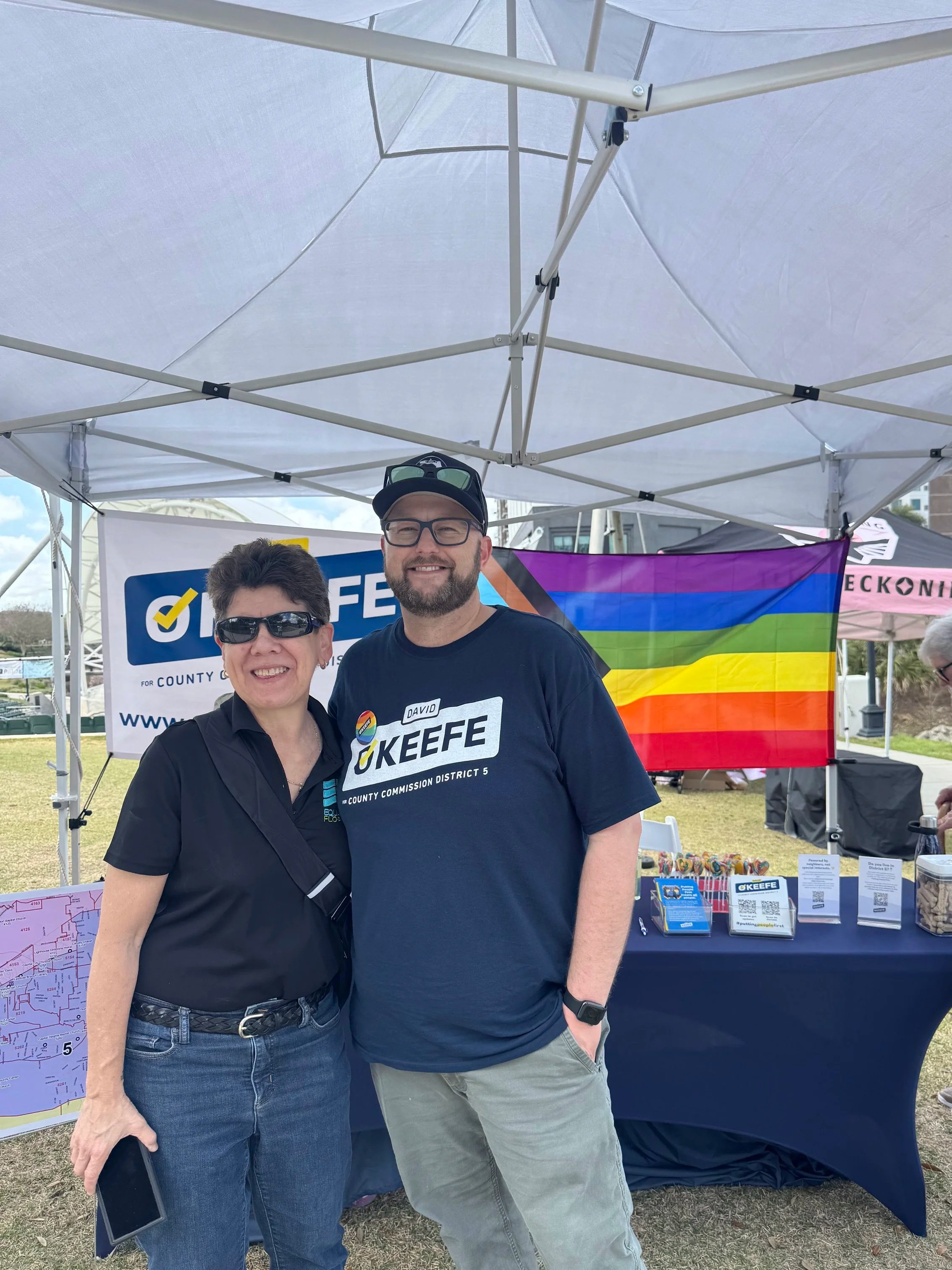 Two people standing under a white canopy tent at an outdoor event. The person on the left is wearing sunglasses and a black shirt, while the person on the right has a beard, glasses on a cap, and a black T-shirt with campaign-related text. Behind them are various promotional materials, a rainbow pride flag, and a sign supporting a county commissioner election.