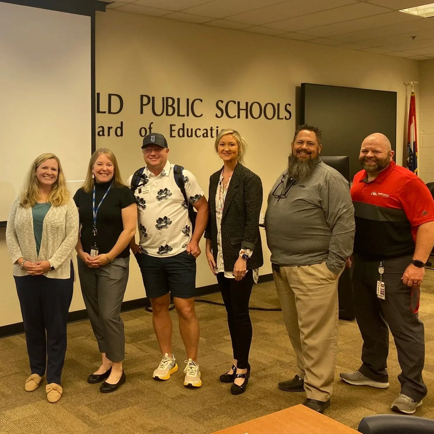 A group of six adults standing in a room with a wall behind them that has text about public schools and education. They are smiling and posing for the photo.