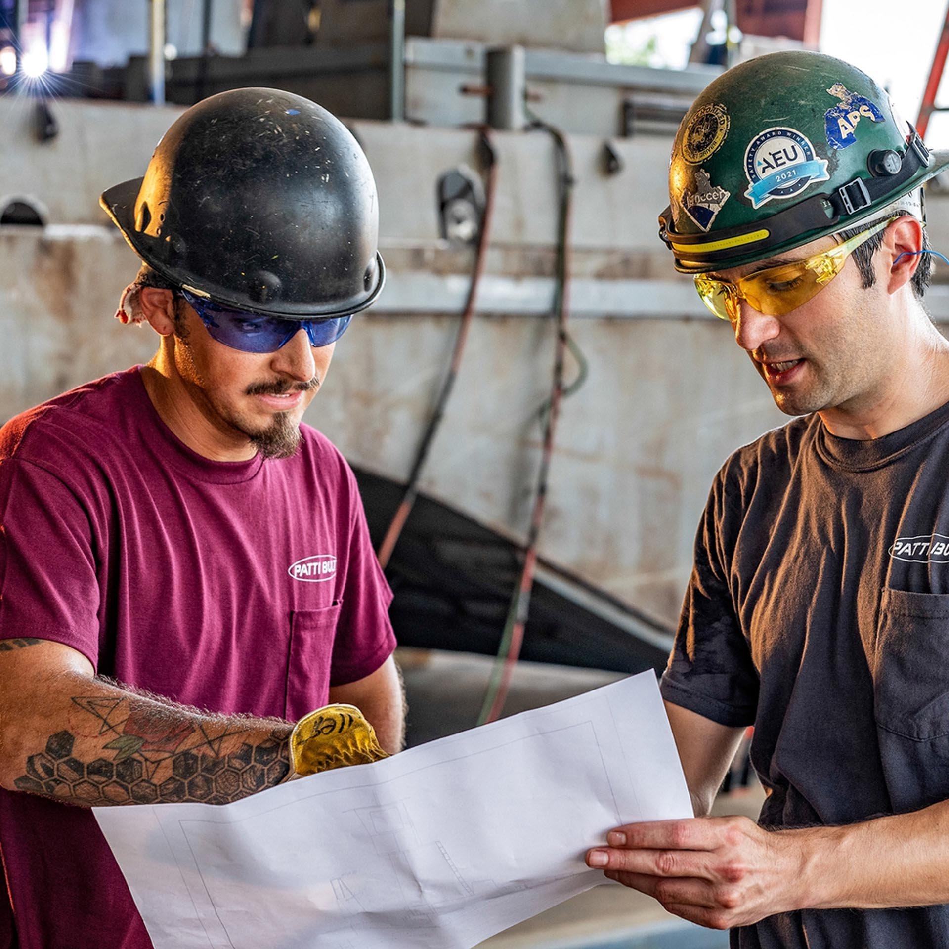 Two construction workers wearing helmets and safety glasses reviewing blueprints together at a construction site.