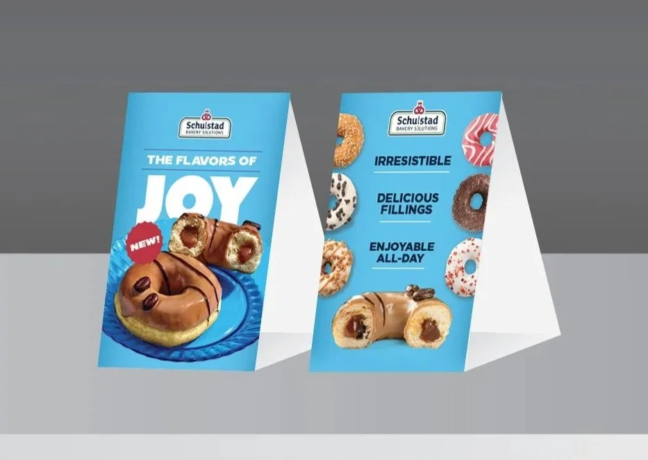 Two promotional standees for Schulstad Bakery Solutions, showcasing donut products with various flavors and fillings. The left standee emphasizes joy and features donuts with chocolate and coffee beans, labeled as new. The right standee highlights ir