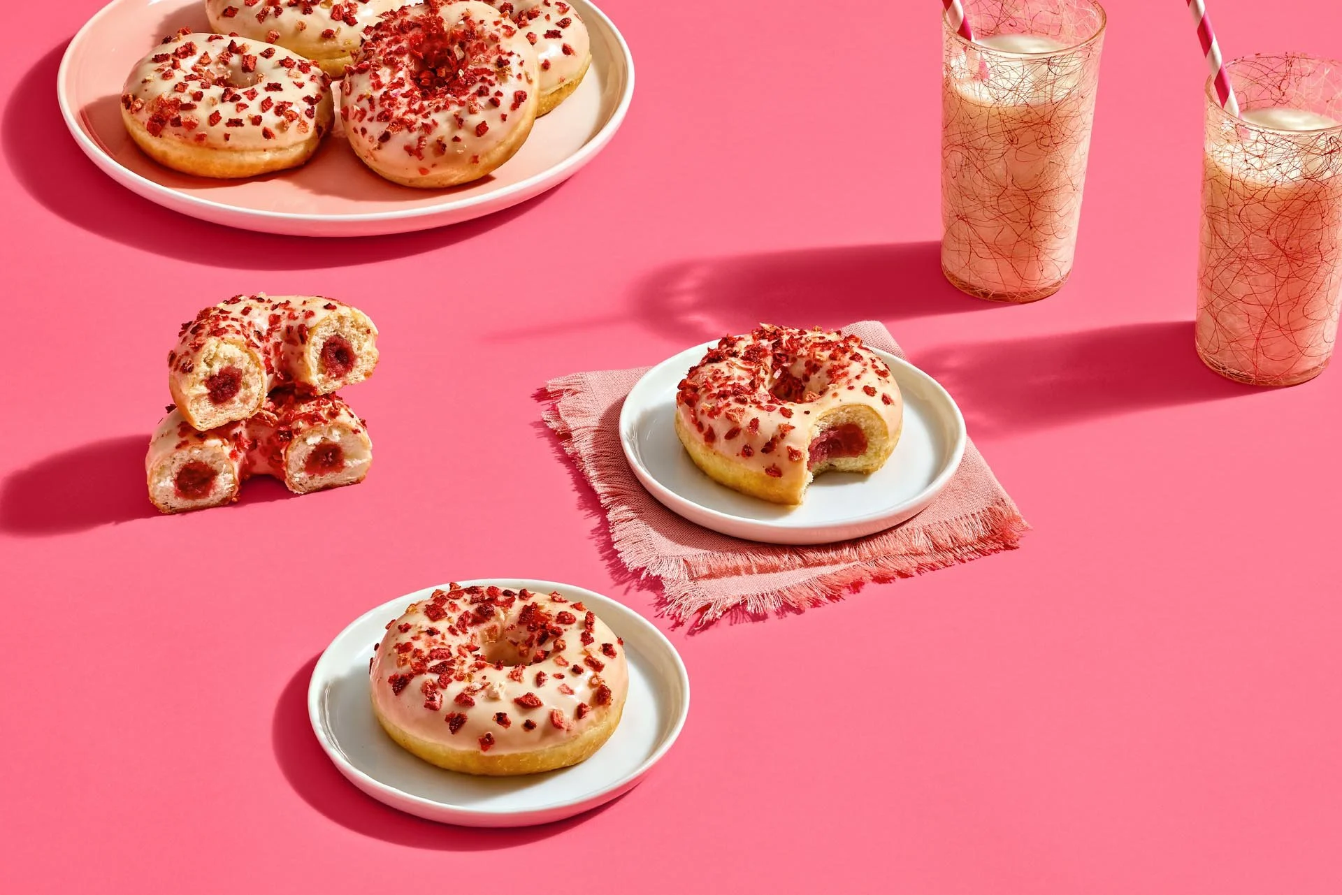 Pink background with white plates holding glazed donuts topped with red sprinkles, some donuts are cut in half showing red filling inside. Two pink and gold patterned glasses with straws are also on the table.