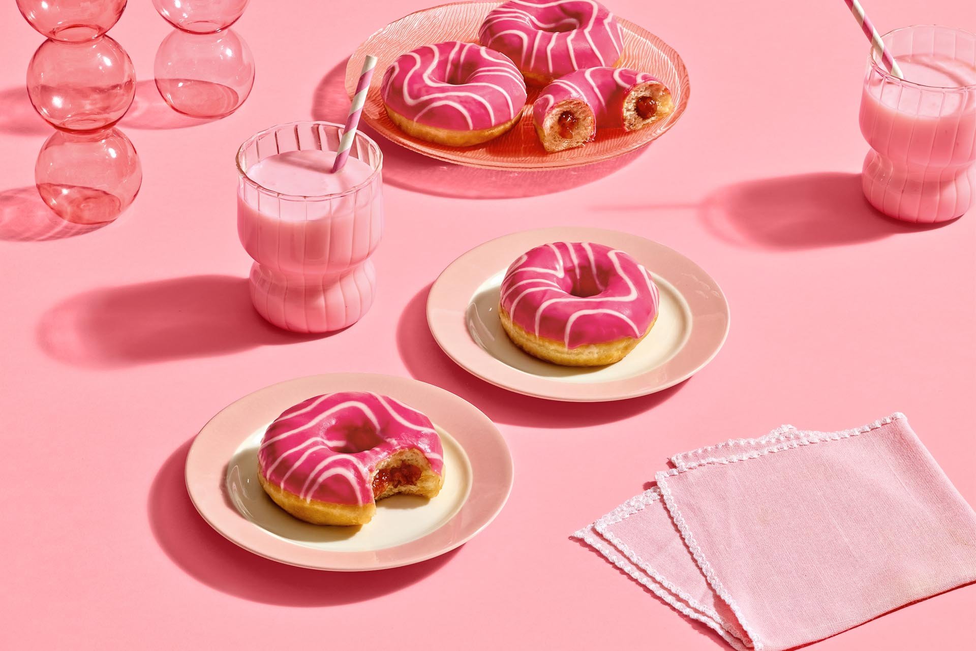Pink background table with pink frosted donuts topped with white icing on small pink plates. Two glasses of pink milk with striped straws. Pink napkin with scalloped edges.