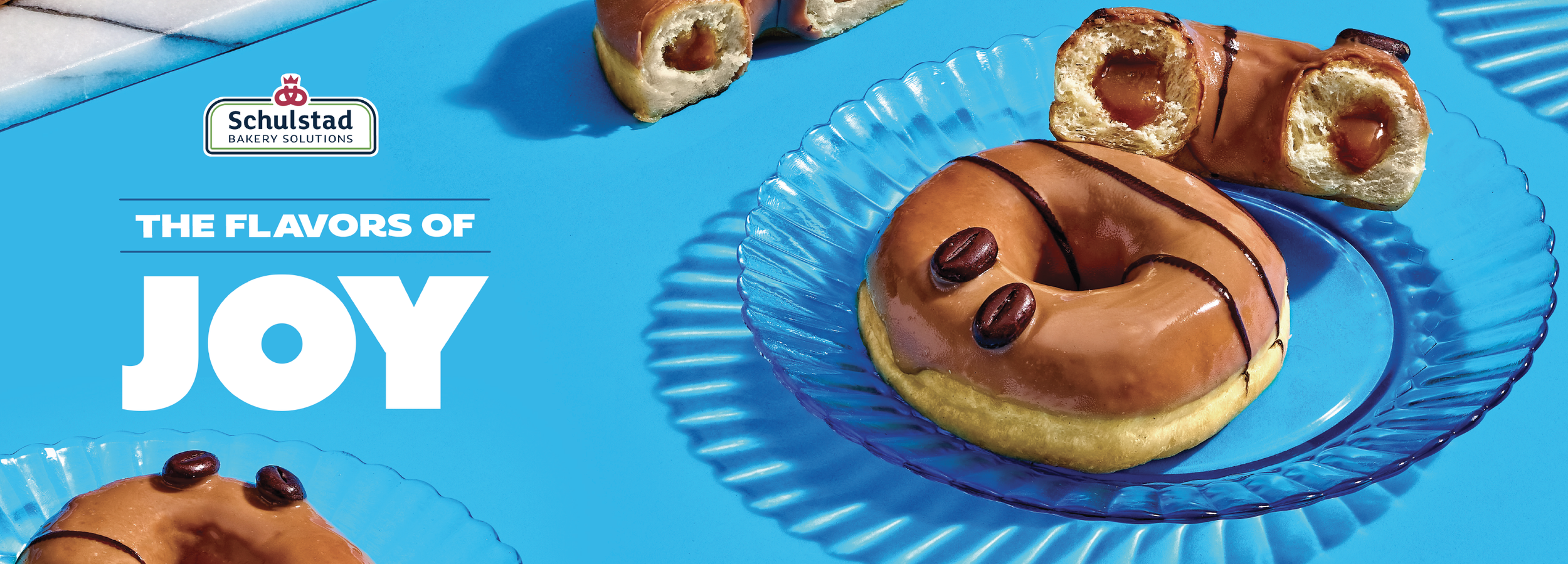 Decorative baked good in the shape of a coffee bean with coffee beans on top, on a blue glass plate, on a bright blue background, with text "The Flavors of Joy" and a Bakery Solutions logo.