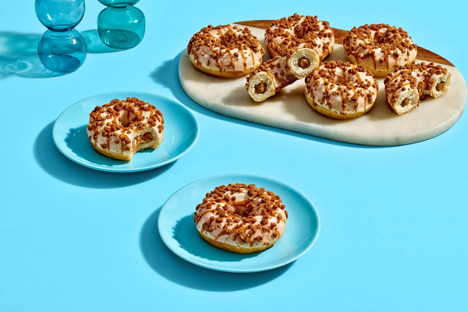Assorted donuts with pink glaze and brown crumbs on blue plates, on a blue background with blue glass vases.