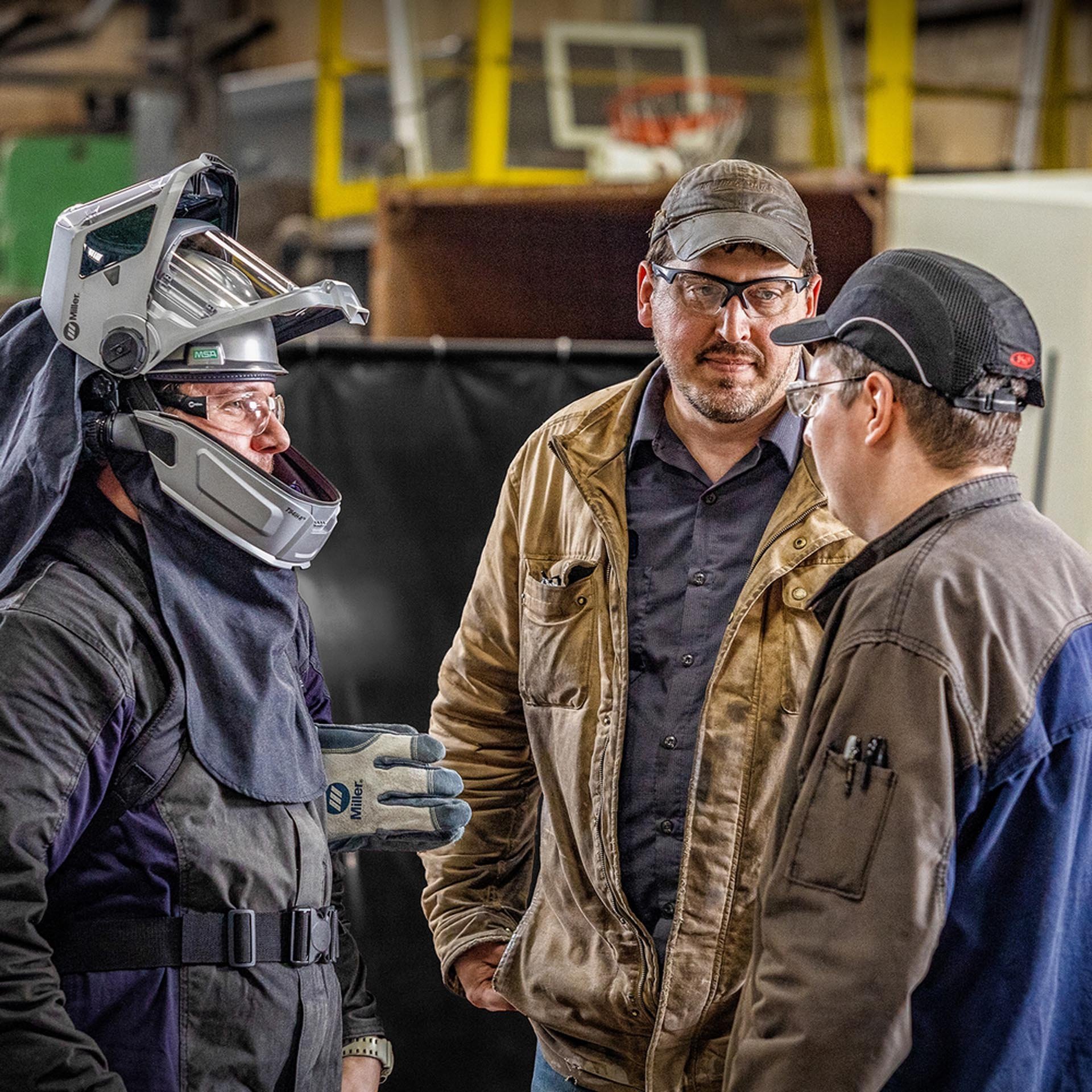Three men in a workshop or warehouse having a conversation. One man, dressed in work gear, is wearing a helmet, gloves, and safety glasses. The second man is wearing a cap and glasses, and the third man, in a brown jacket, has a beard and is listenin