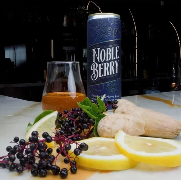A can of Noble Berry, a glass of berry soda, ginger root, lemon slices, blackberry clusters, lime wedge, and fresh mint on a table.