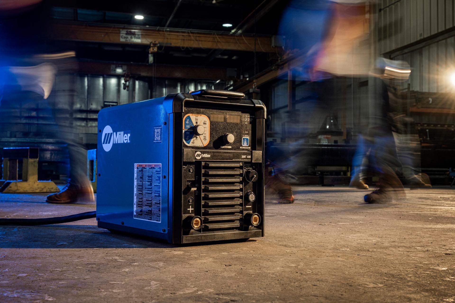 A blue Miller-branded welding machine on a factory floor, with blurred motion of workers walking past in the background at night.